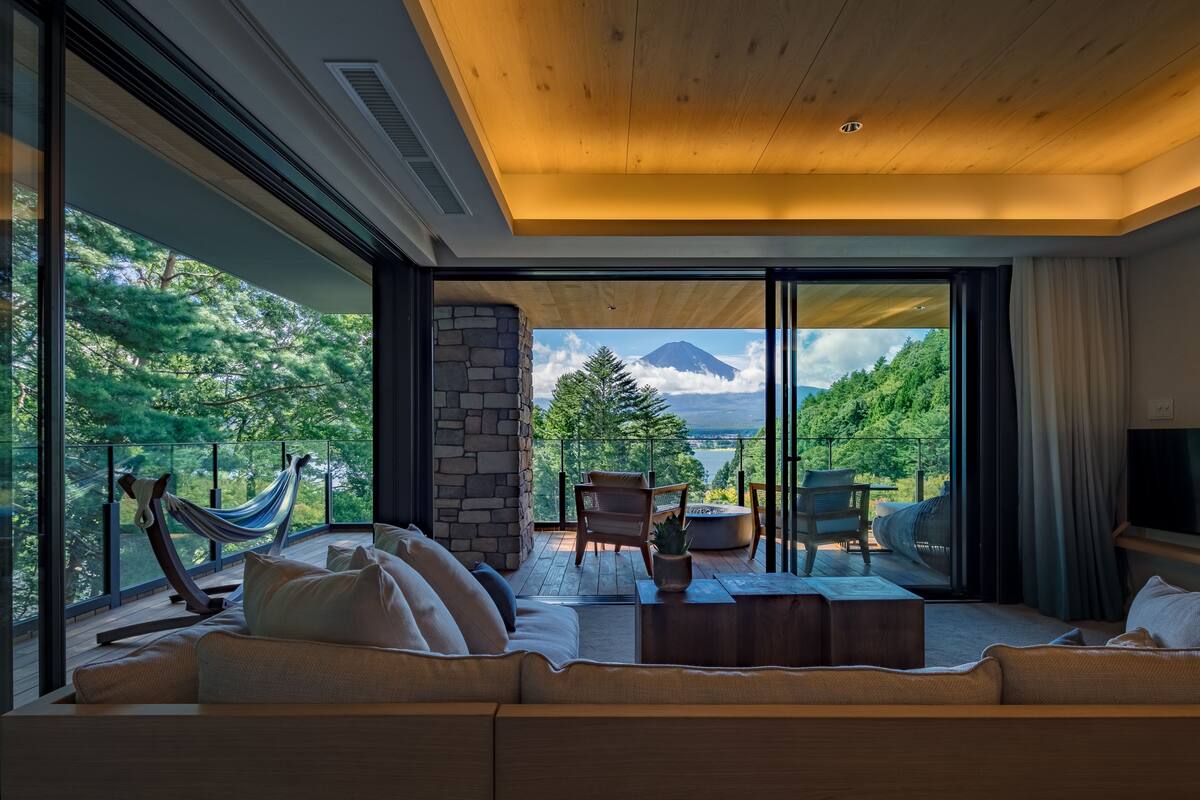 A modern living room with a large sofa, a coffee table, and a hammock. It features a stone pillar, a fireplace, and a view of a mountain through large glass doors.