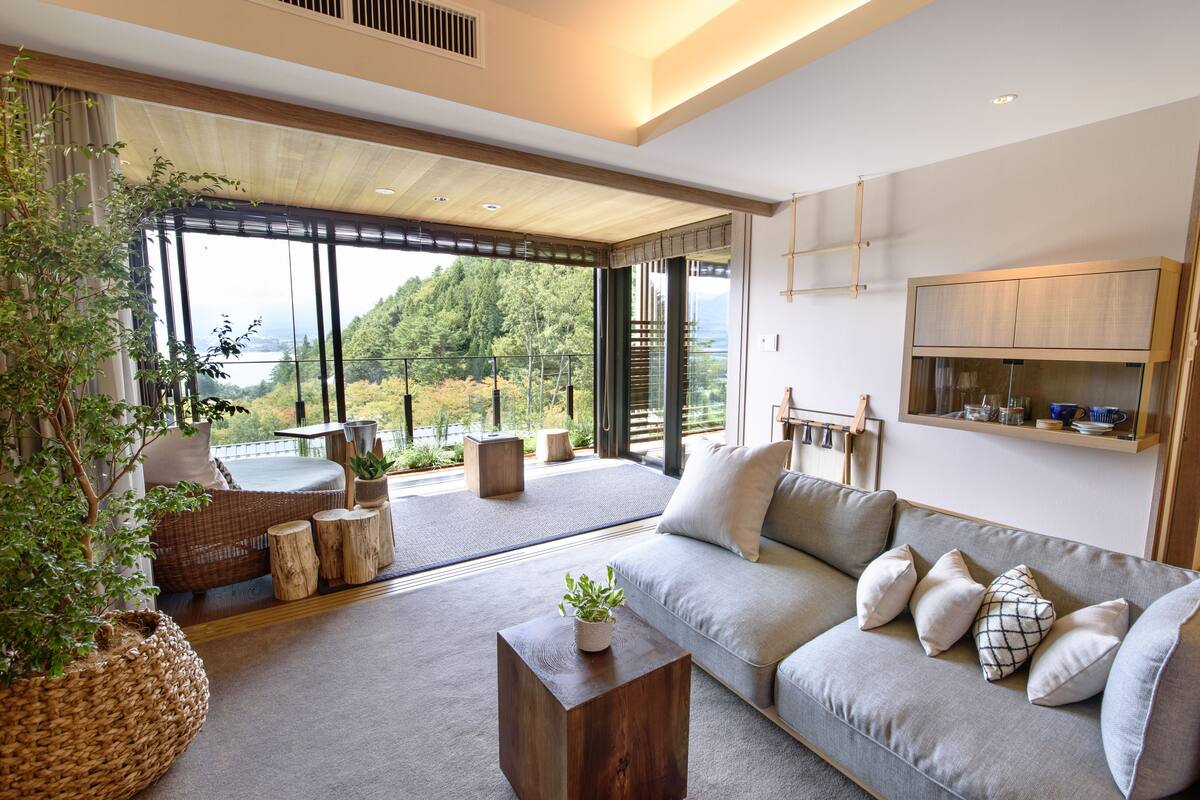 A modern living room with a large sofa, a coffee table, and a view of a forested area through large windows.