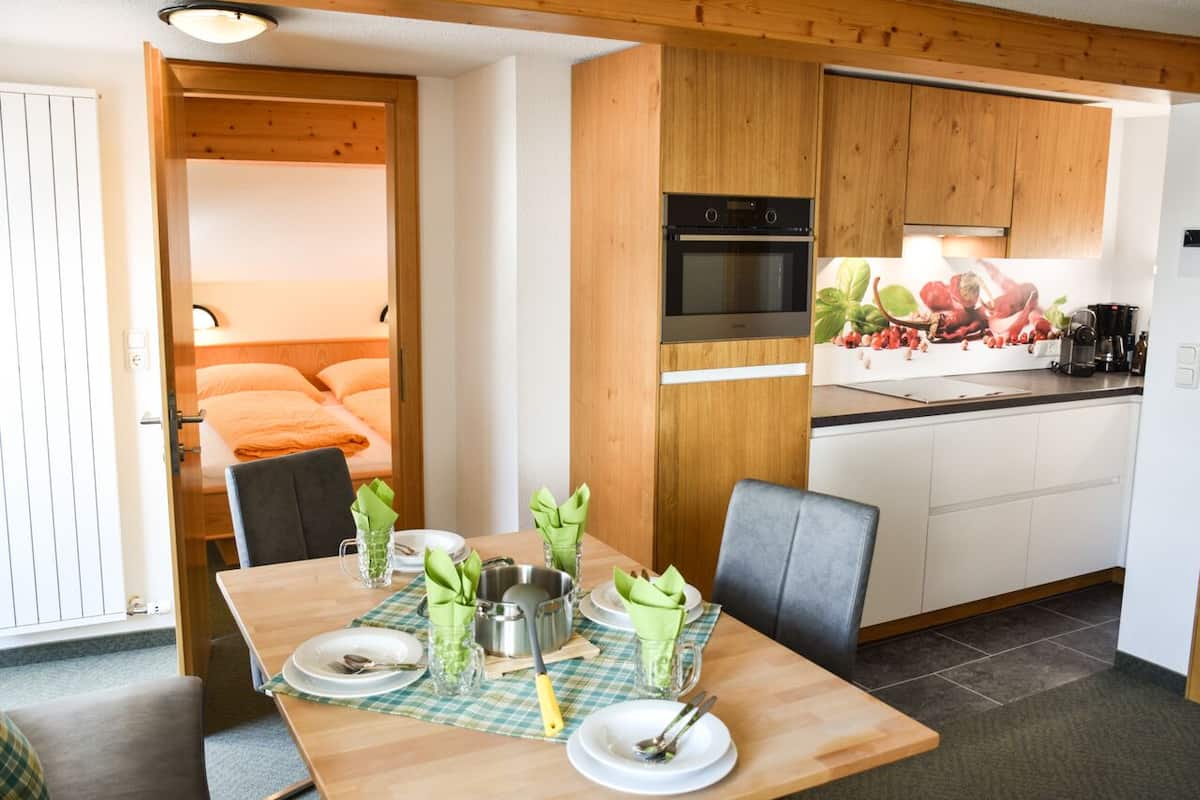 Habitación de hotel compacta con una mesa de comedor de madera dispuesta para dos personas, una kitchenette con estufa y horno, y una cama con ropa de cama naranja.