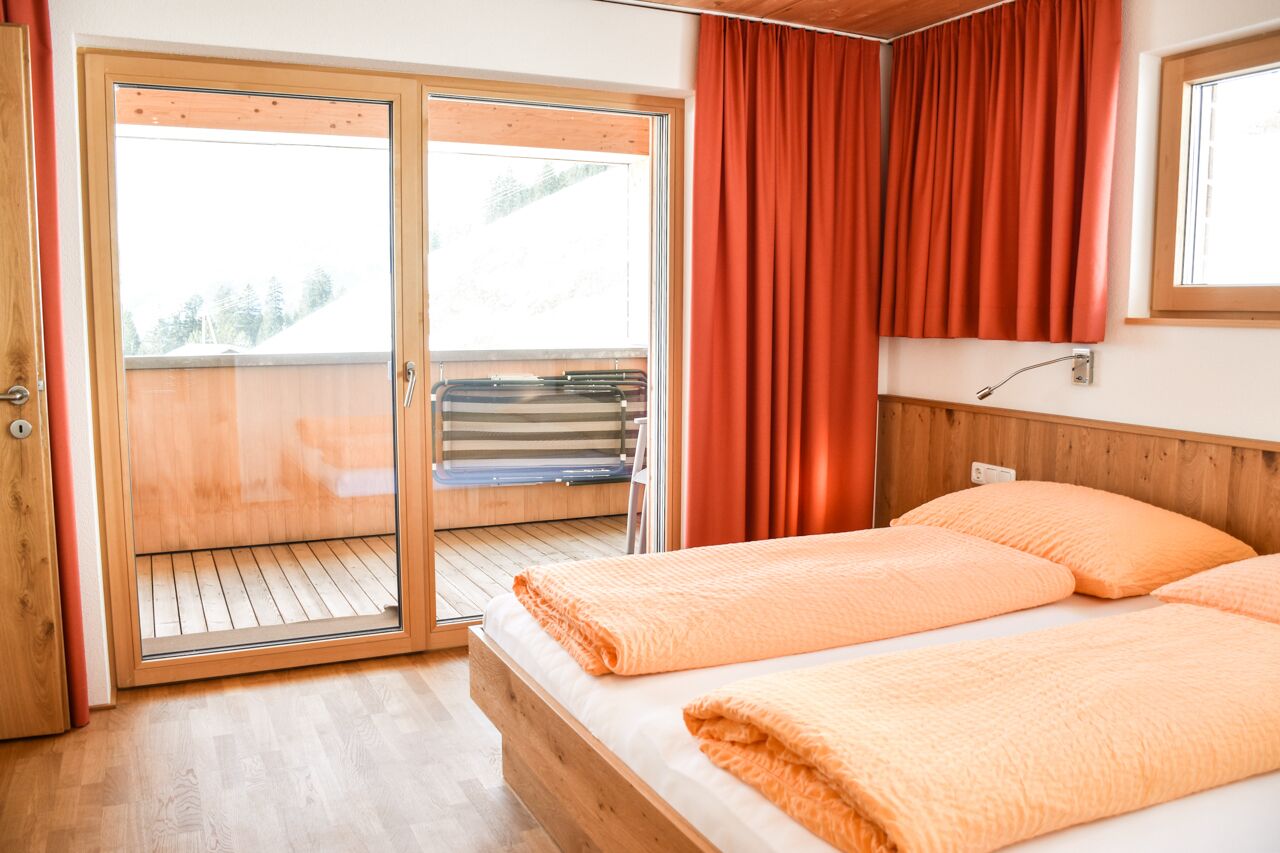 Une chambre à coucher avec un lit en bois, une literie orange, une porte-fenêtre coulissante donnant sur un balcon et un rideau rouge.