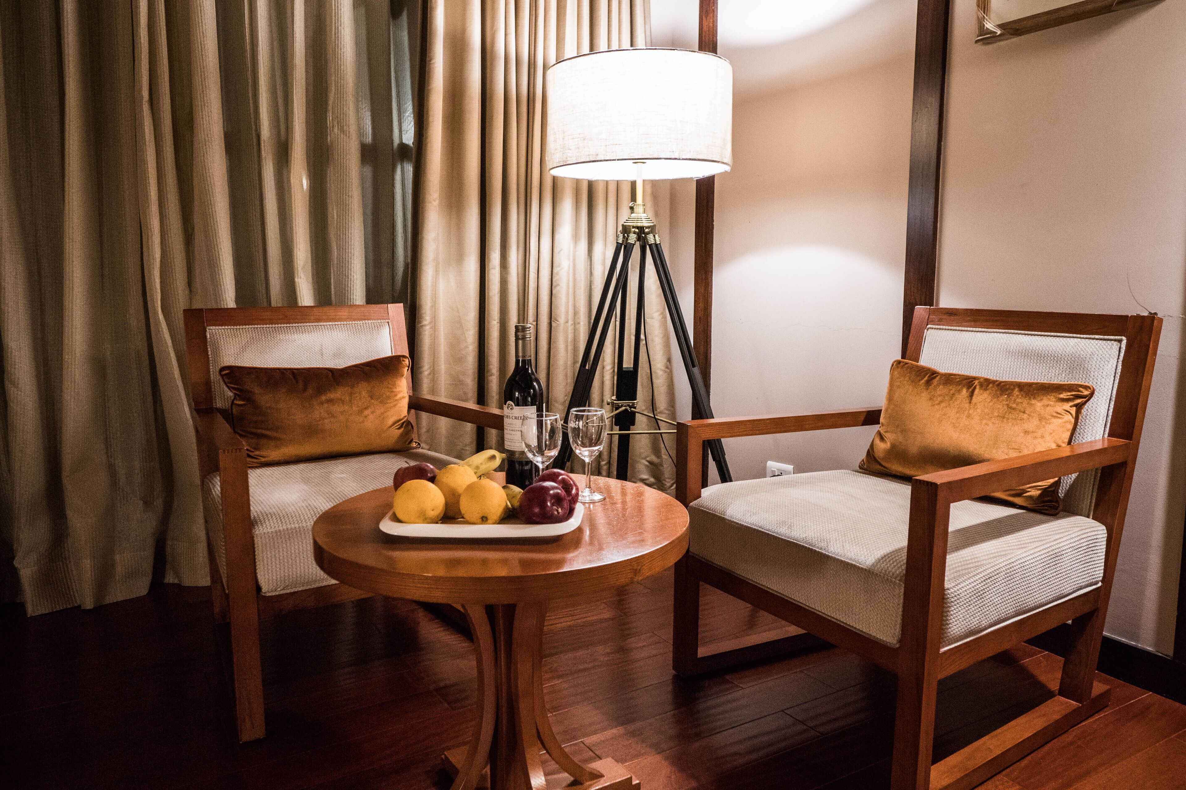 Two wooden chairs with cushions, a small round table with fruits and glasses, and a floor lamp.