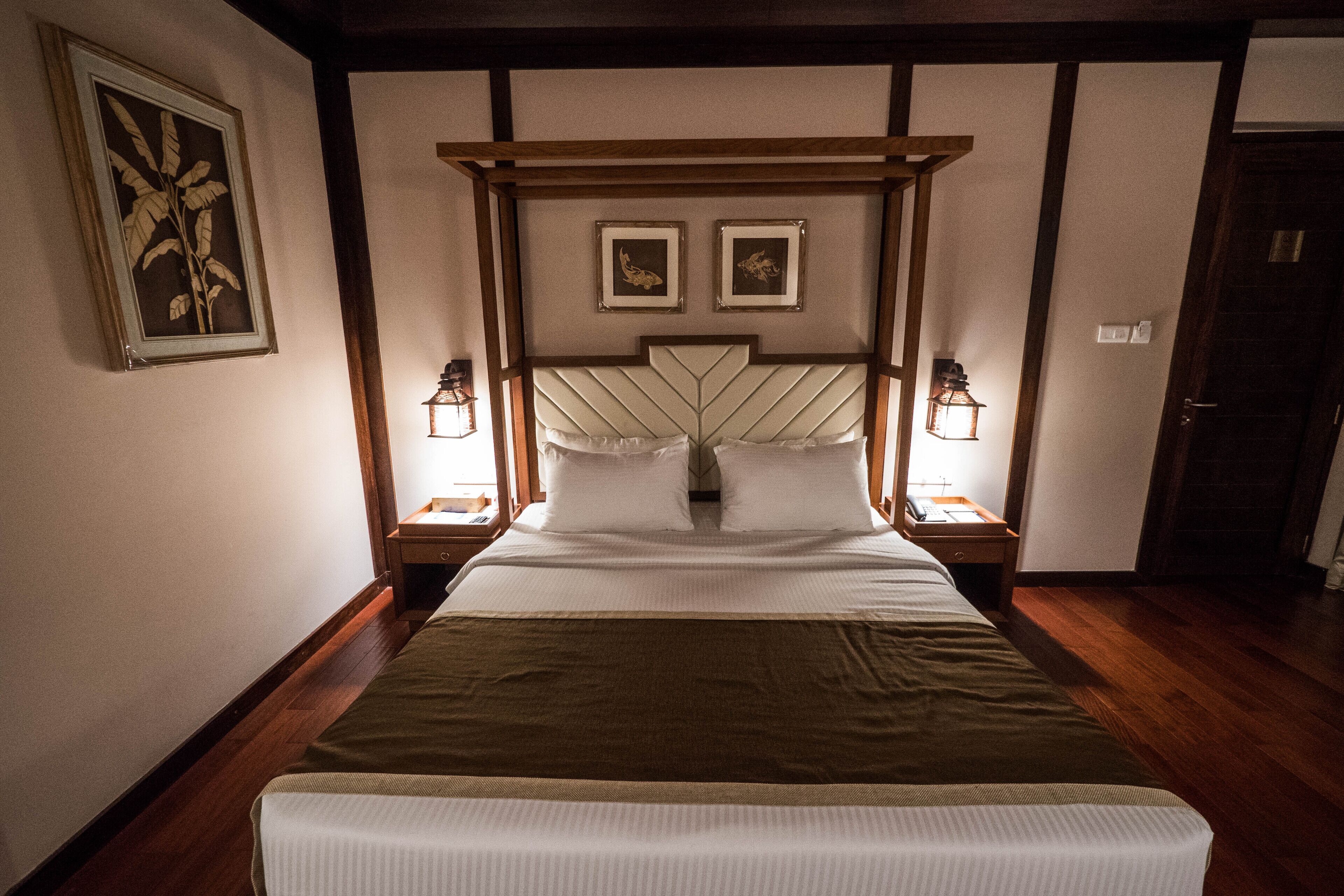Un lit bien fait, avec une tête de lit en bois, deux tables de chevet avec des lampes et une plante encadrée accrochée au mur.