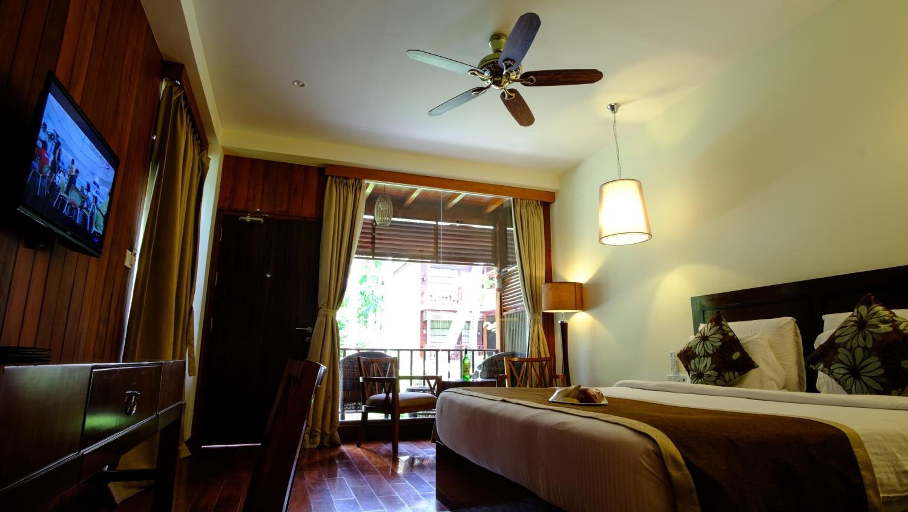 A hotel room with a bed, a desk, a chair, a ceiling fan, a TV, and a window with curtains.