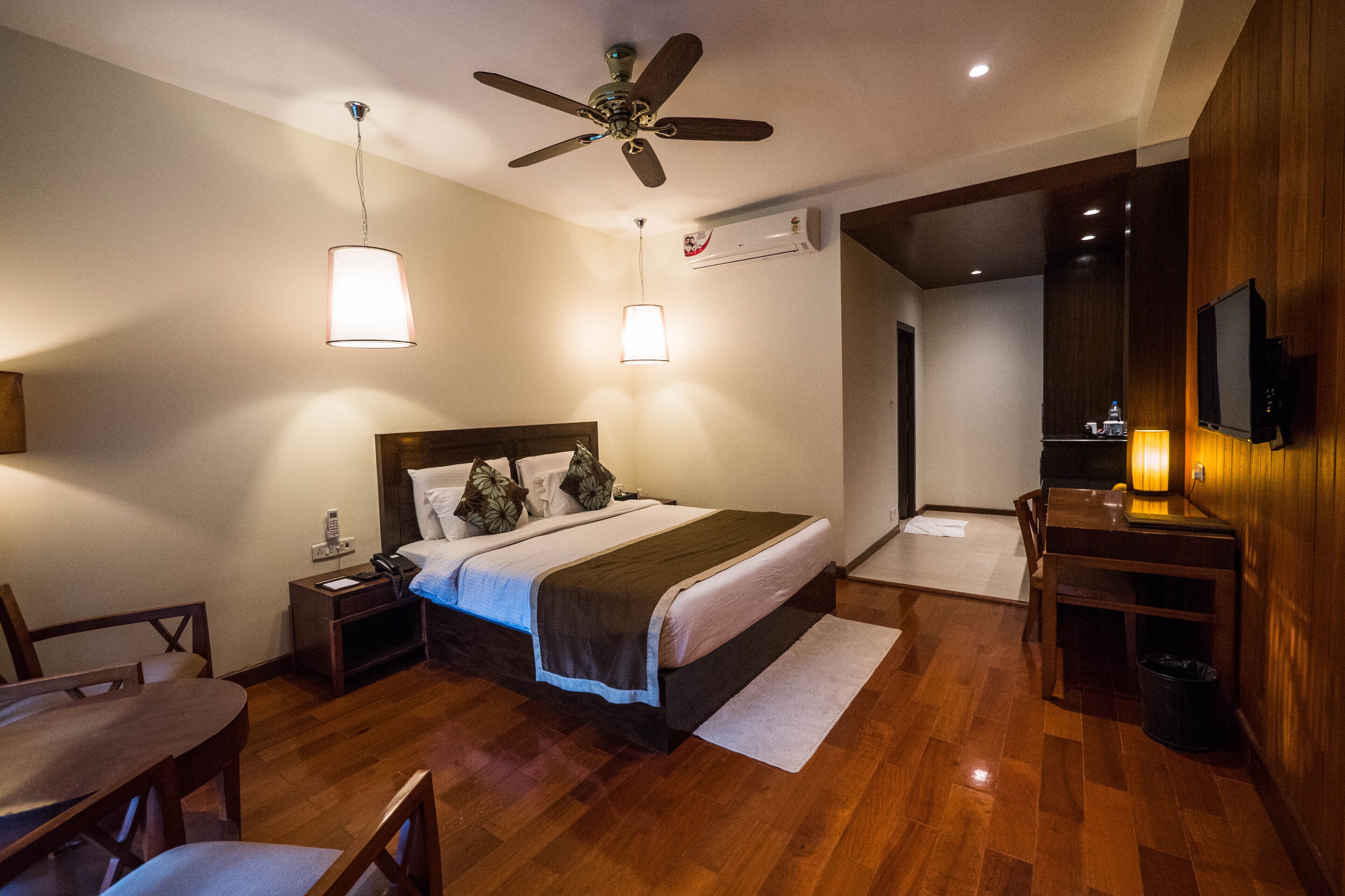 A hotel room with a bed, bedside tables, a desk, a chair, a ceiling fan, and a TV.