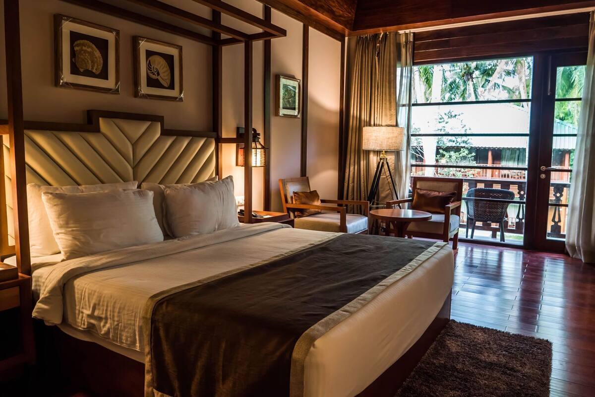 A hotel room with a large bed, a desk, two chairs, a window with curtains, and framed pictures on the wall.