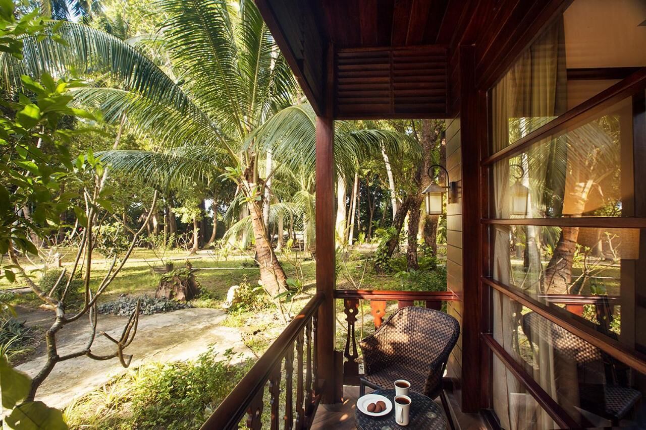 Ein Balkon mit Korbstuhl, einem kleinen Tisch mit Teller und zwei Tassen, und Blick auf einen tropischen Garten mit Palmen.