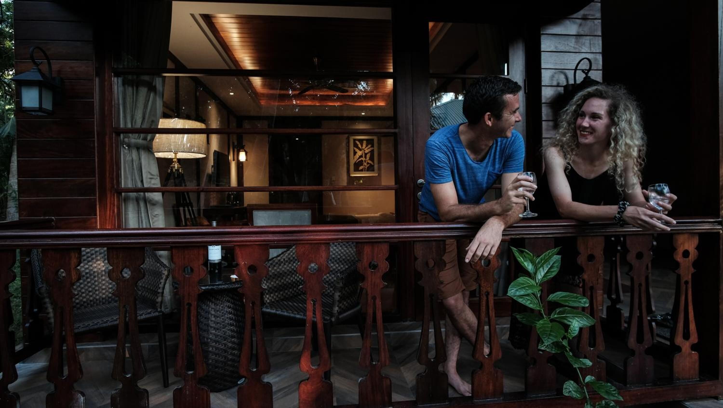 A man and a woman sitting on a wooden balcony railing, holding glasses, with a view of an indoor dining area and a plant in the foreground.