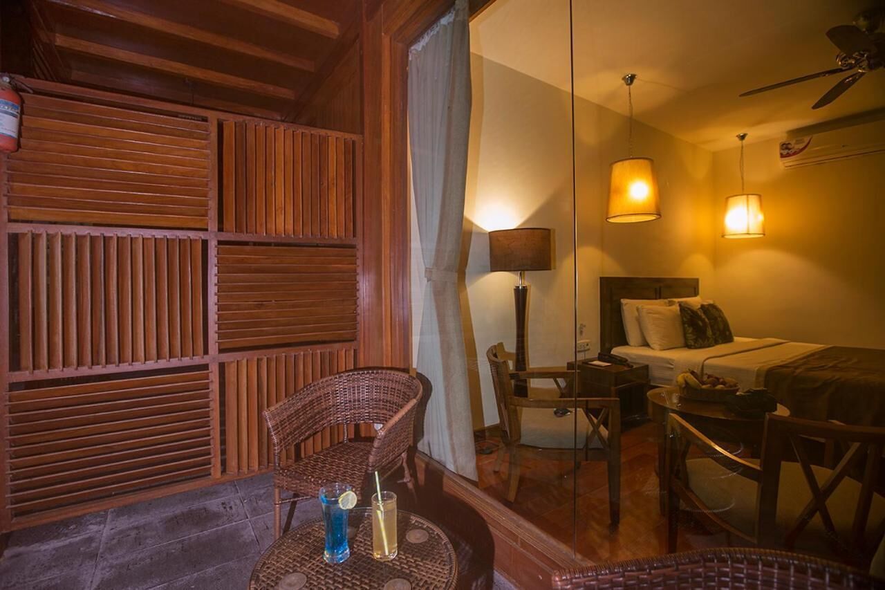 A cozy room with a bed, a dining table, and a wicker chair. There is a window with blinds, a lamp, and a ceiling fan.