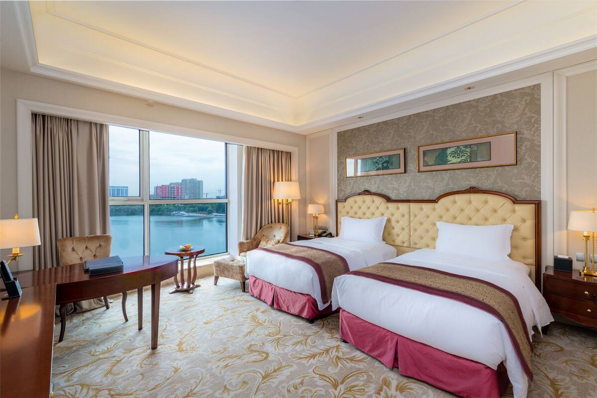 A hotel room with two beds, a desk, and a chair. There are two lamps on the nightstands and a view of a river and buildings through the window.