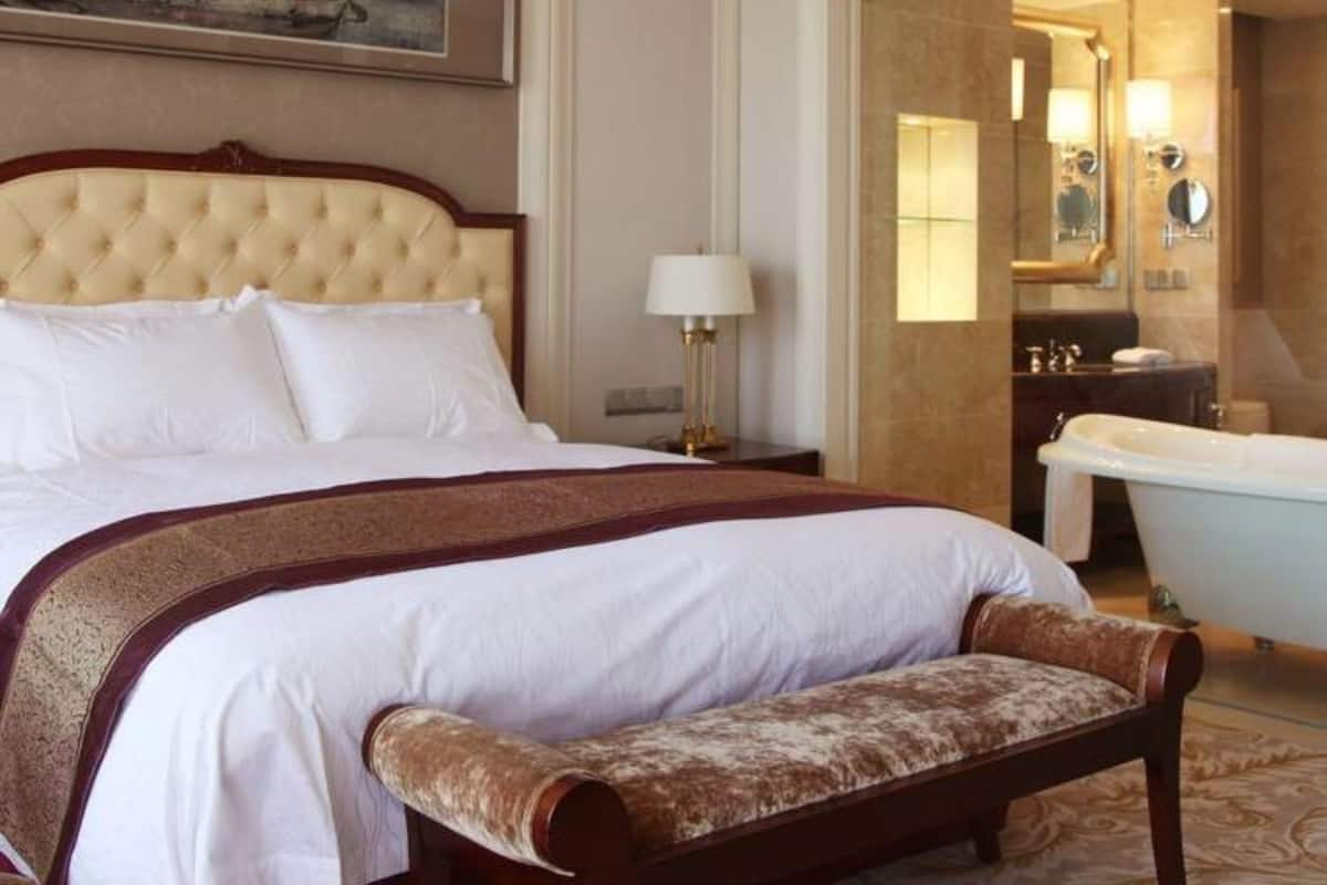 A hotel room with a large bed, a bench, a nightstand, a lamp, a painting, and a bathroom with a bathtub and mirror.