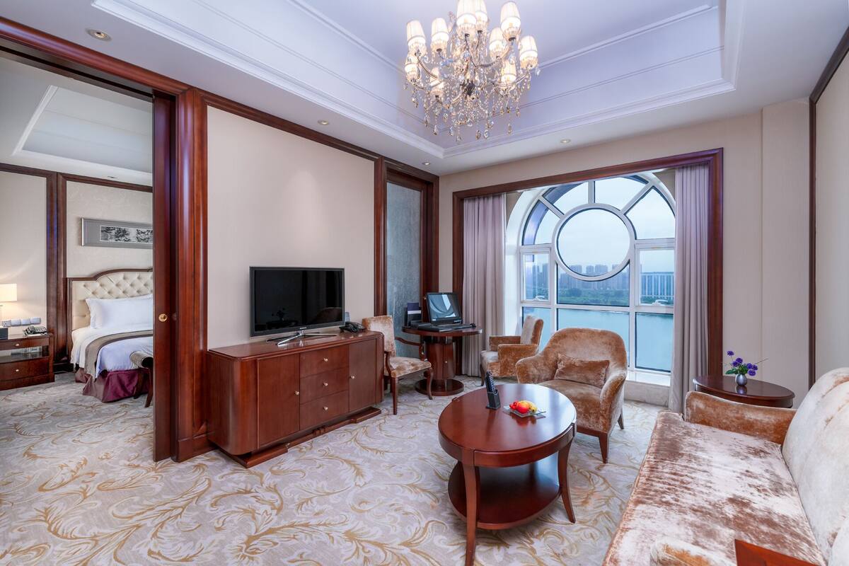 A hotel room with a large bed, a TV, a desk with a laptop, a sofa, and a large window with a view of the city.