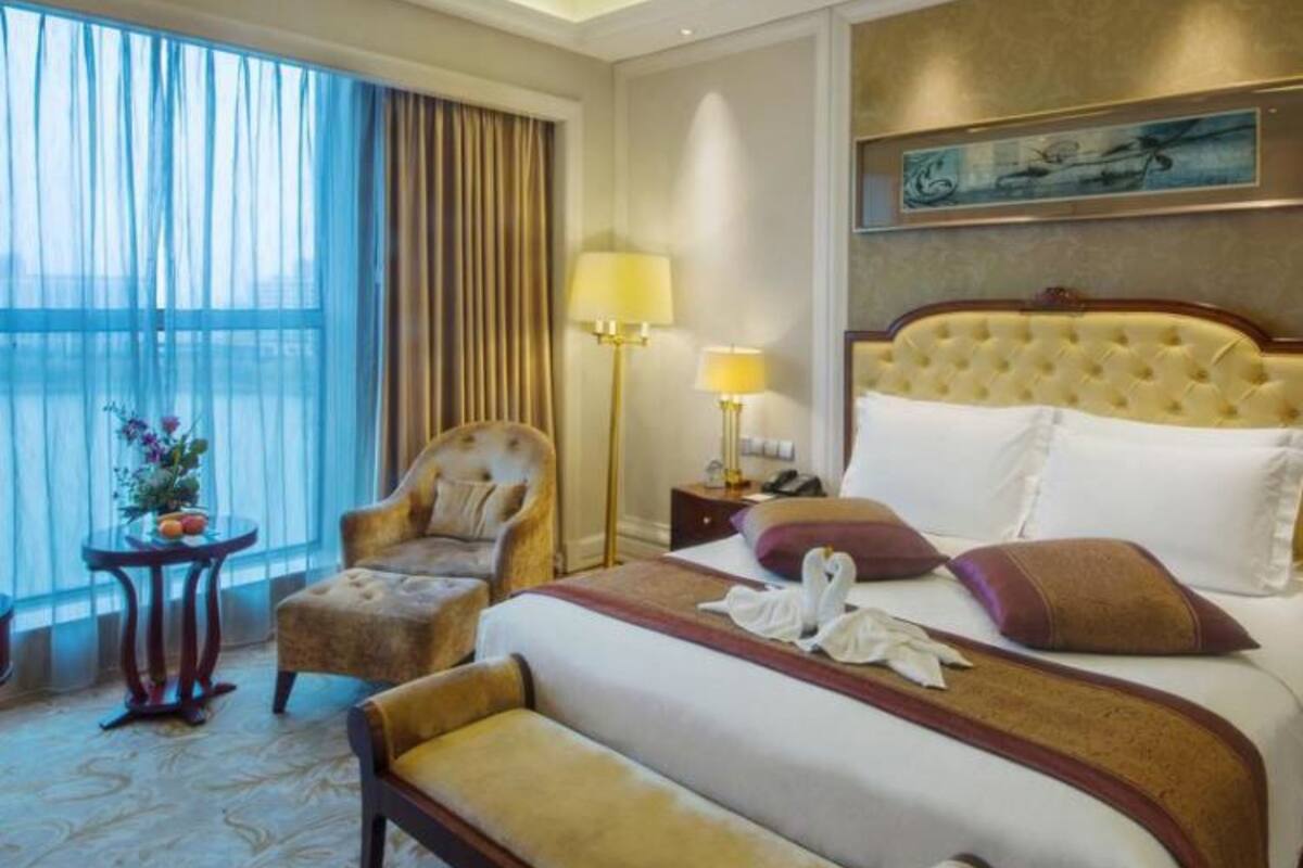 A hotel room with a large bed, a chair, a bedside table with lamps, a painting on the wall, and a view through the window.
