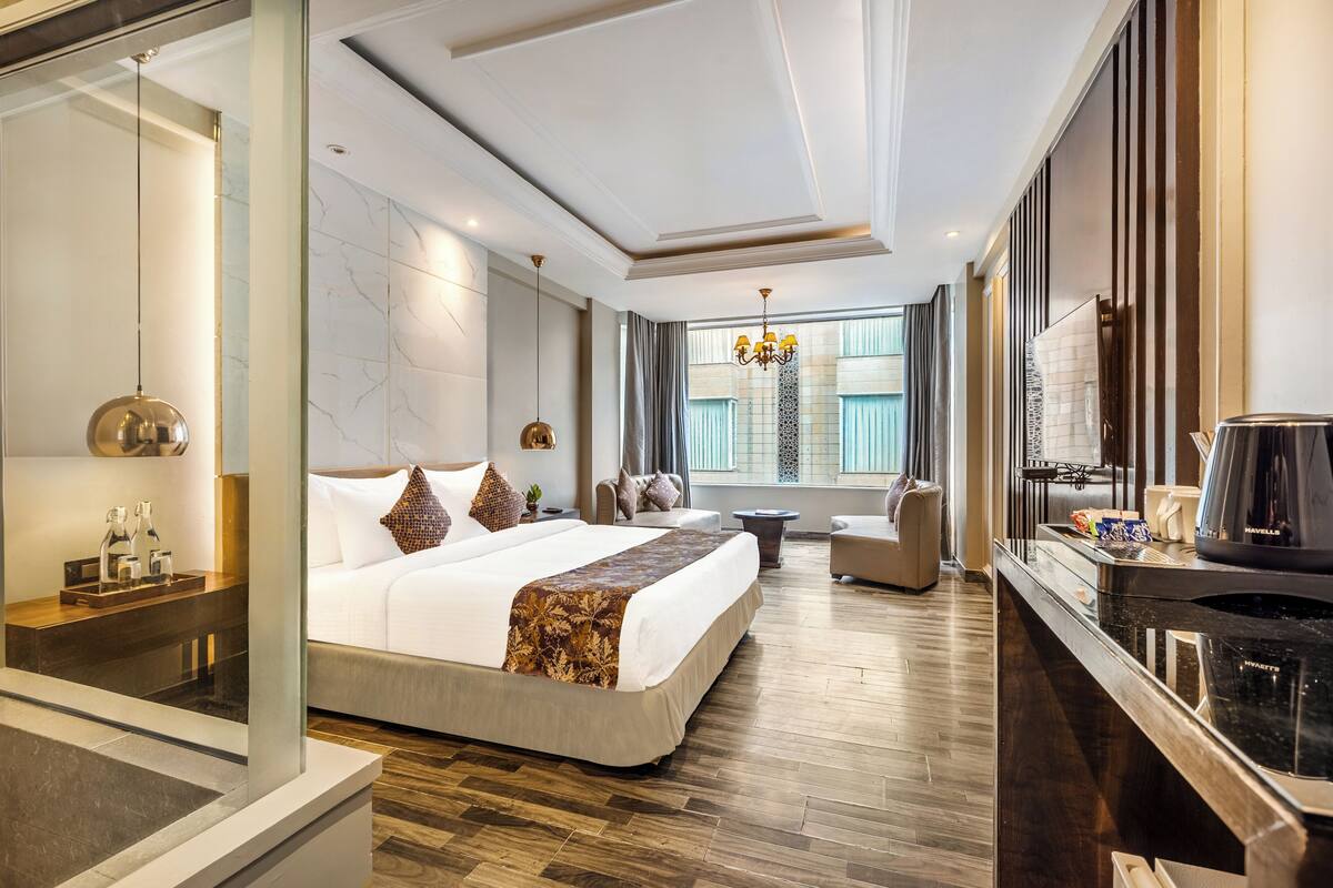 A modern hotel room with a large bed, a sitting area, a coffee station, and a view of the city.