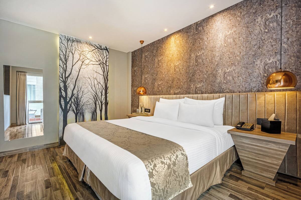 A modern hotel room with a large bed, a wooden floor, a textured wall, and a nightstand with a lamp and phone.