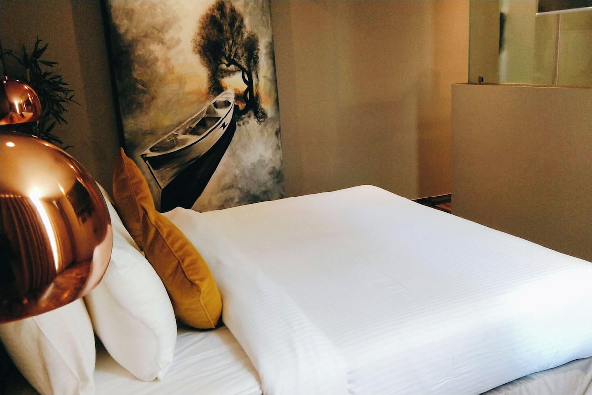 A neatly made bed with white bedding, a single yellow pillow, and a copper-colored lamp on the left.
