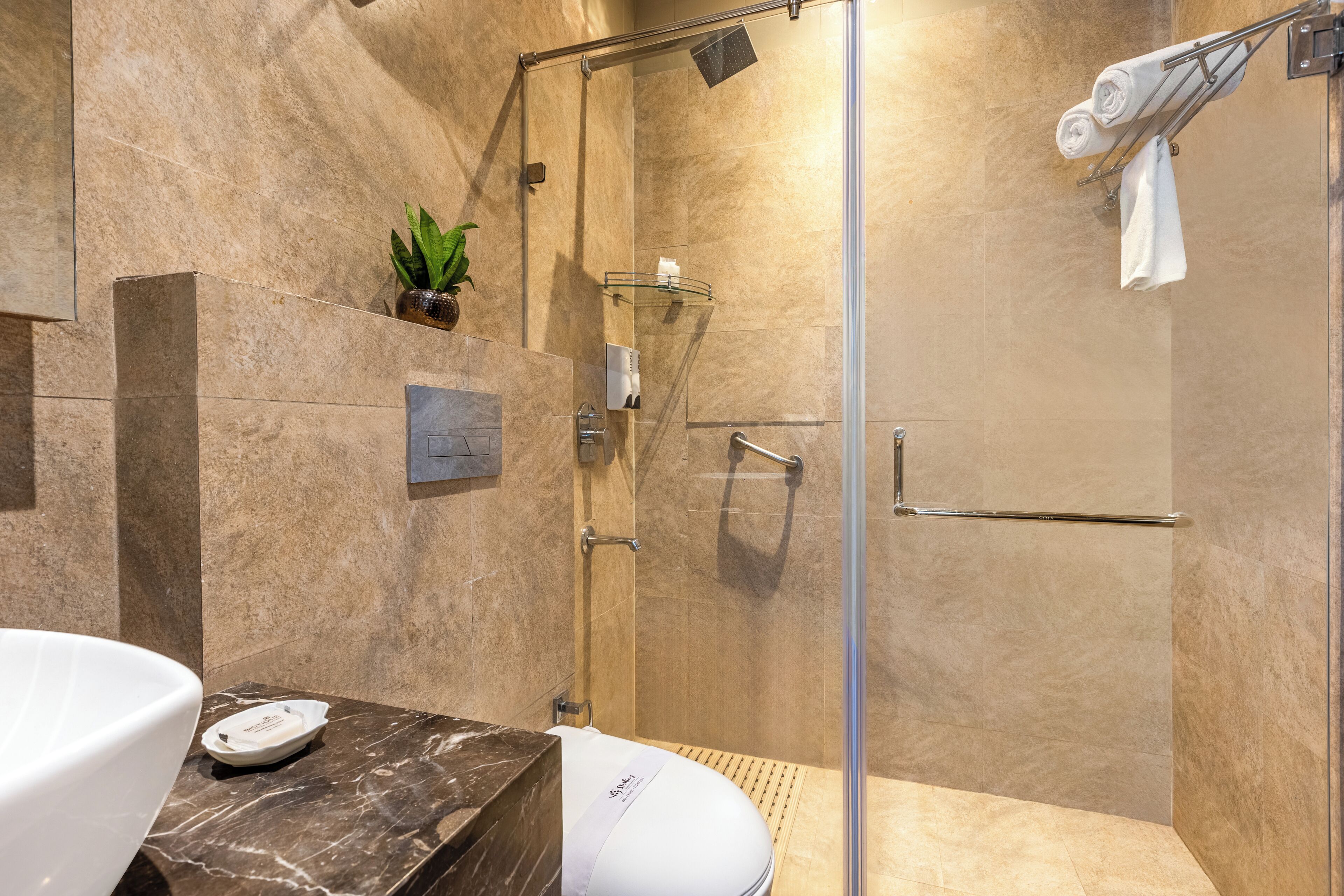 Un baño moderno con ducha de vidrio, encimeras de mármol, inodoro blanco y una pequeña planta en maceta.