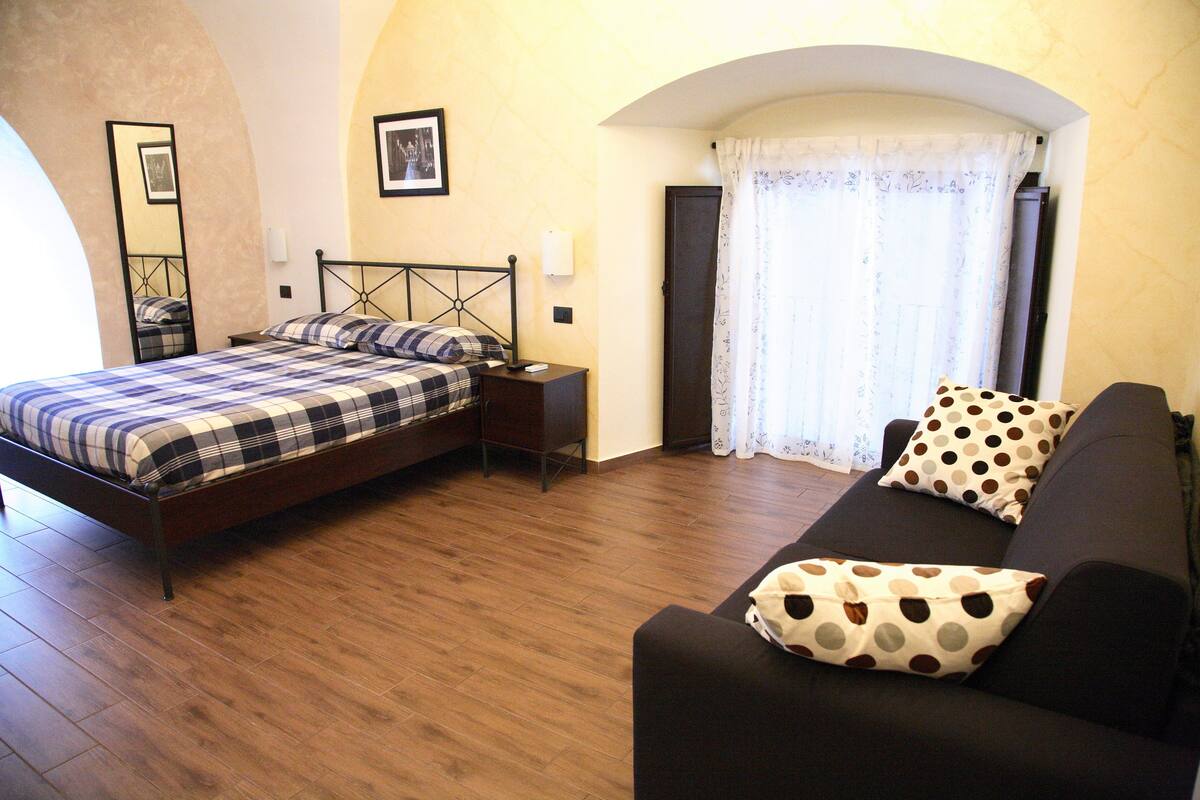 A bedroom with a bed, a nightstand, a sofa, and a window with curtains.