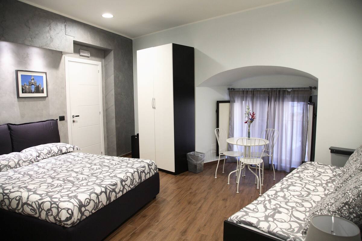 A hotel room with a bed, a sofa, a small table with chairs, a trash bin, and a door leading to an outdoor area.