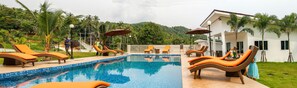Zona de piscina con sillones de descanso y sombrillas, rodeada de exuberante vegetación y una colina al fondo.