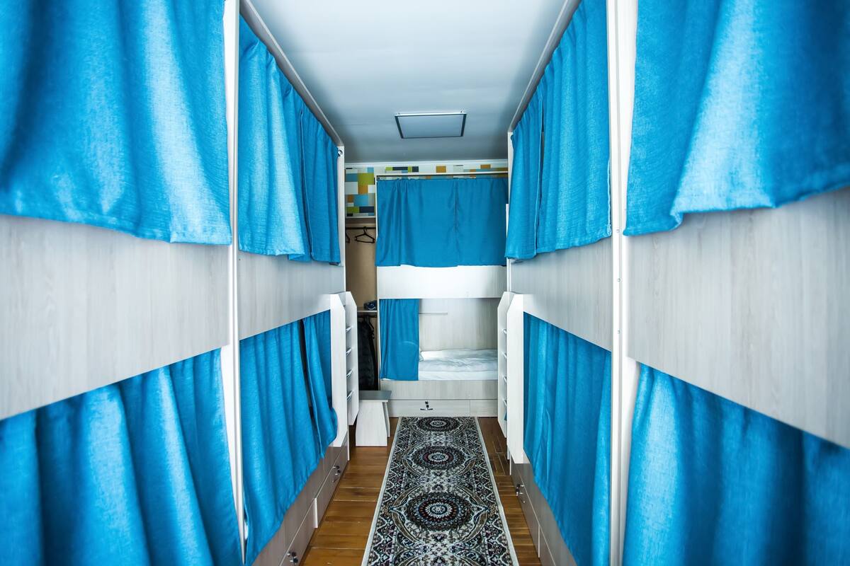 Un pasillo estrecho con literas y cortinas azules, que da a un baño con un tapete estampado.
