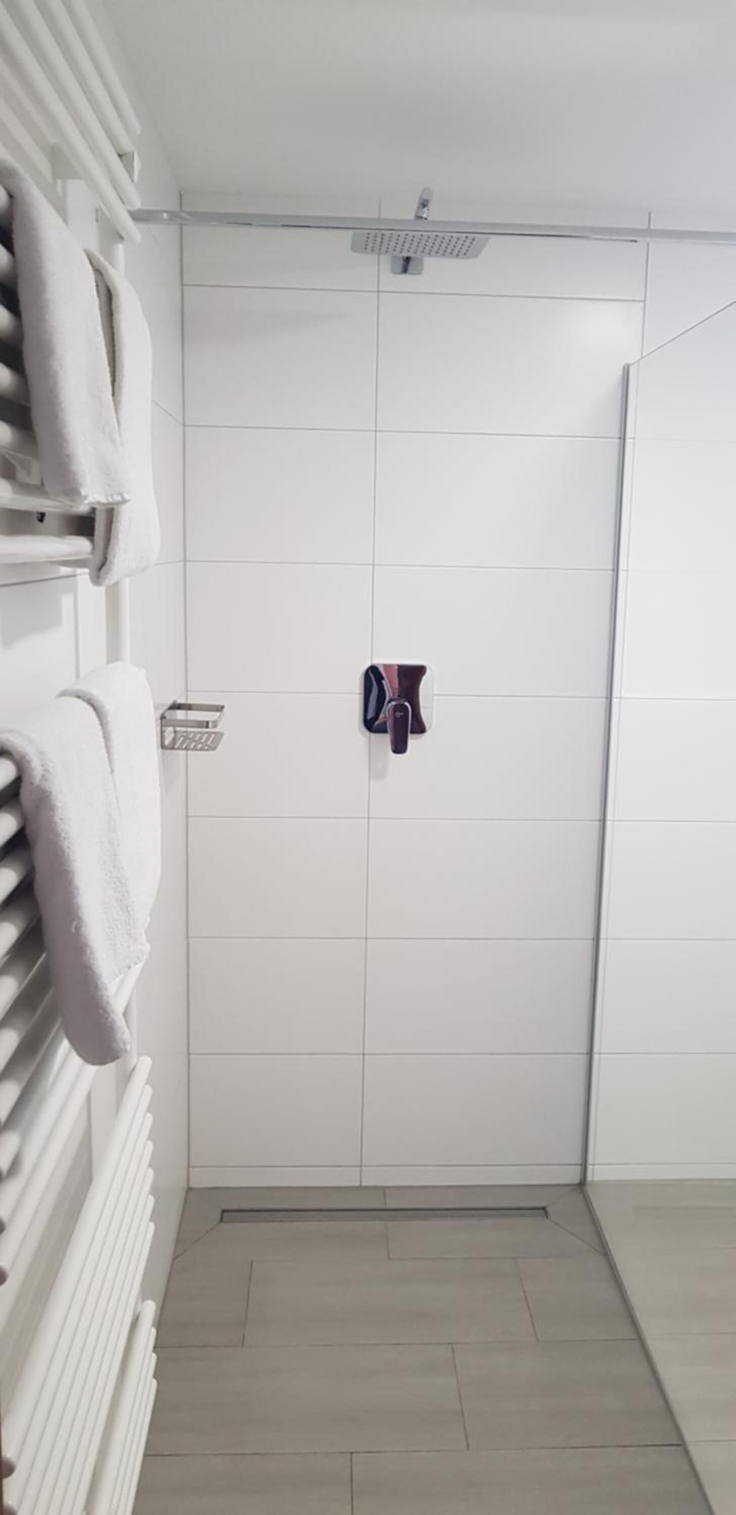 Un baño con paredes de azulejos blancos, zona de ducha y un perchero con toallas.