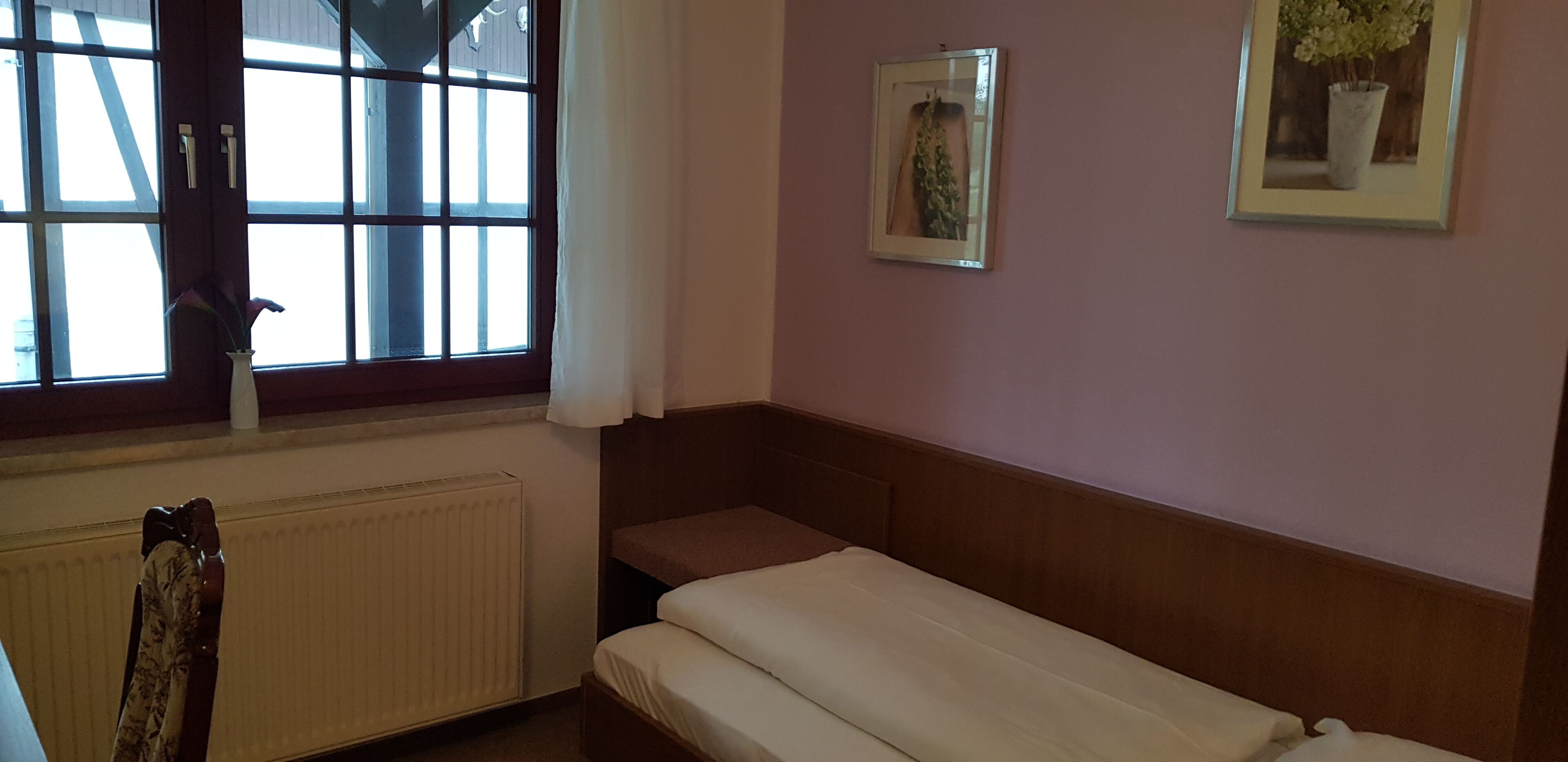 Una habitación pequeña y sencilla con una cama individual, un radiador, una ventana con cortinas y dos cuadros enmarcados en la pared.
