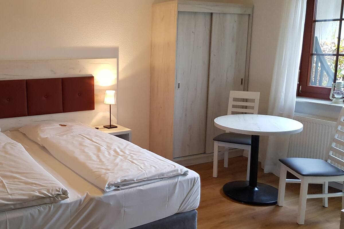 A neatly made bed with white linens, a wooden headboard, a bedside table with a lamp, a small round table with a chair, and a window with curtains.
