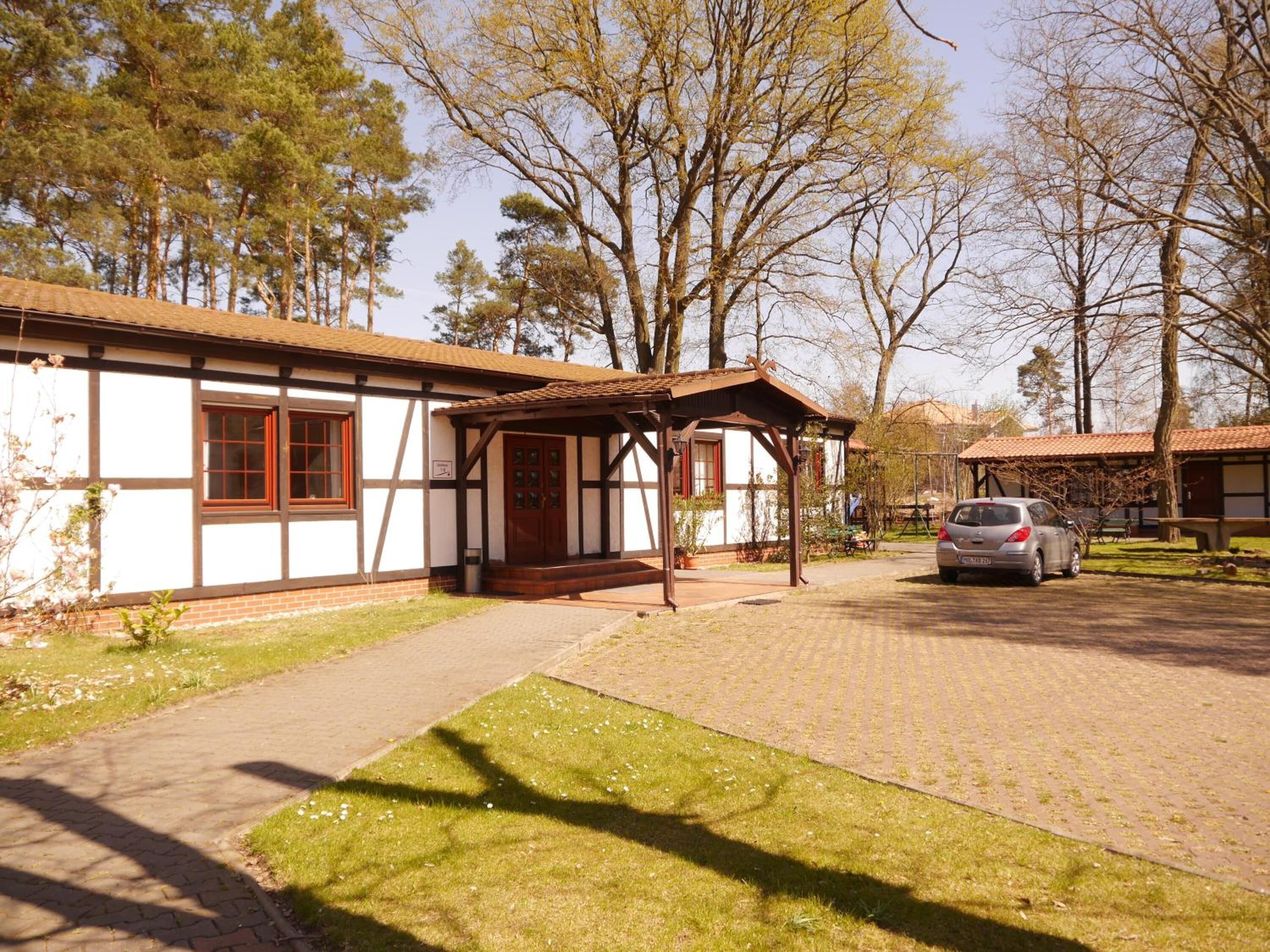 A traditional building with a wooden porch, a car parked in front, and a grassy area with trees in the background.