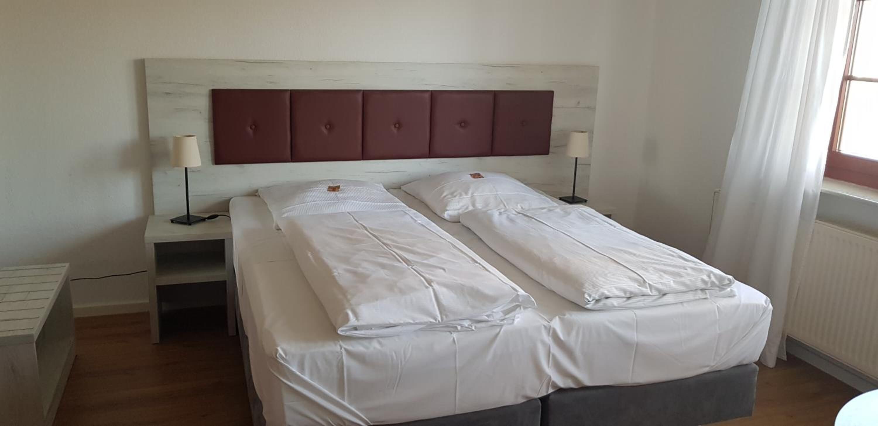 Una cama doble con cabecera de madera, dos mesitas de noche con lámparas y una ventana con cortinas.
