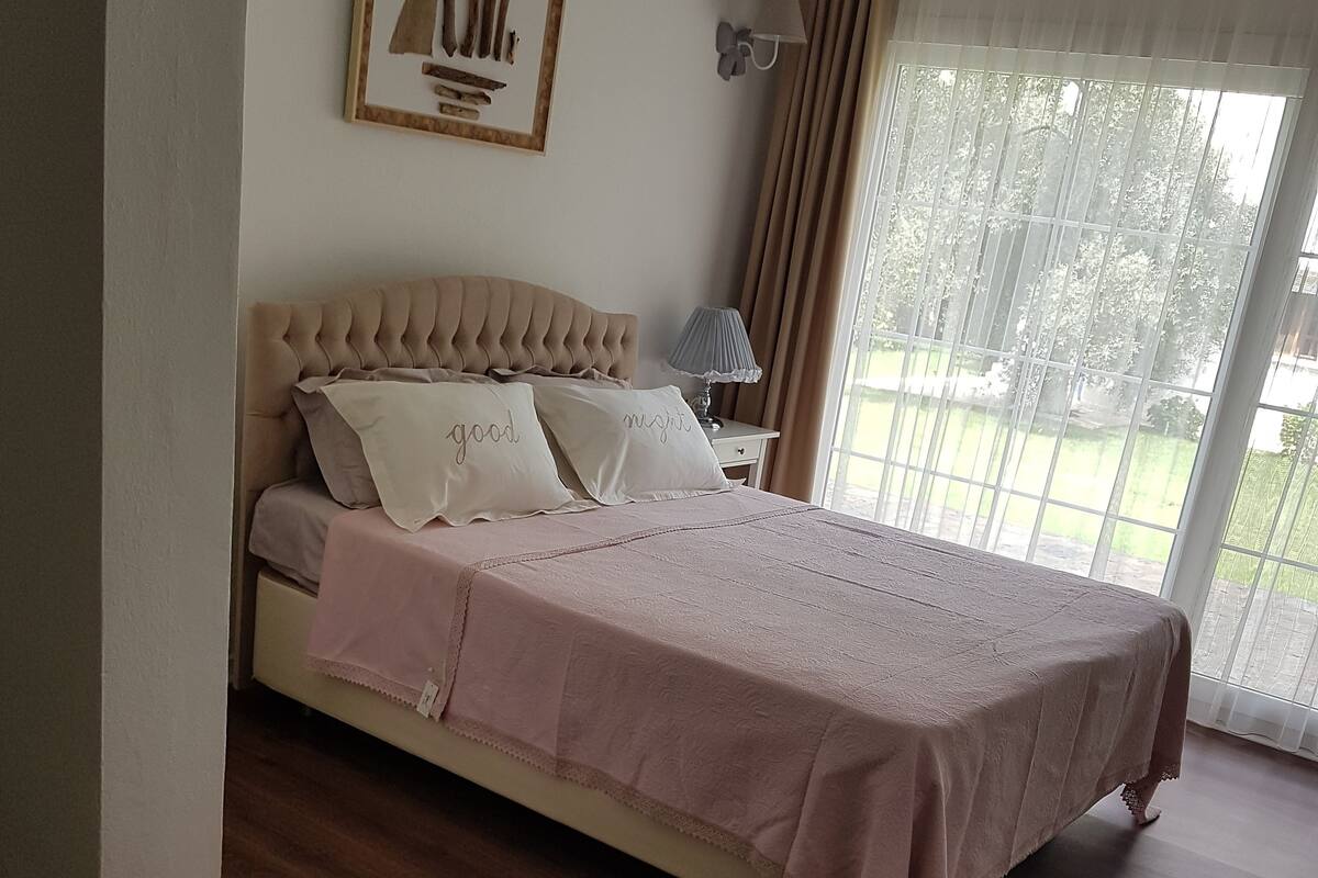 Une chambre à coucher avec un lit, une table de chevet, une lampe, un tableau encadré au mur et une fenêtre avec des rideaux.