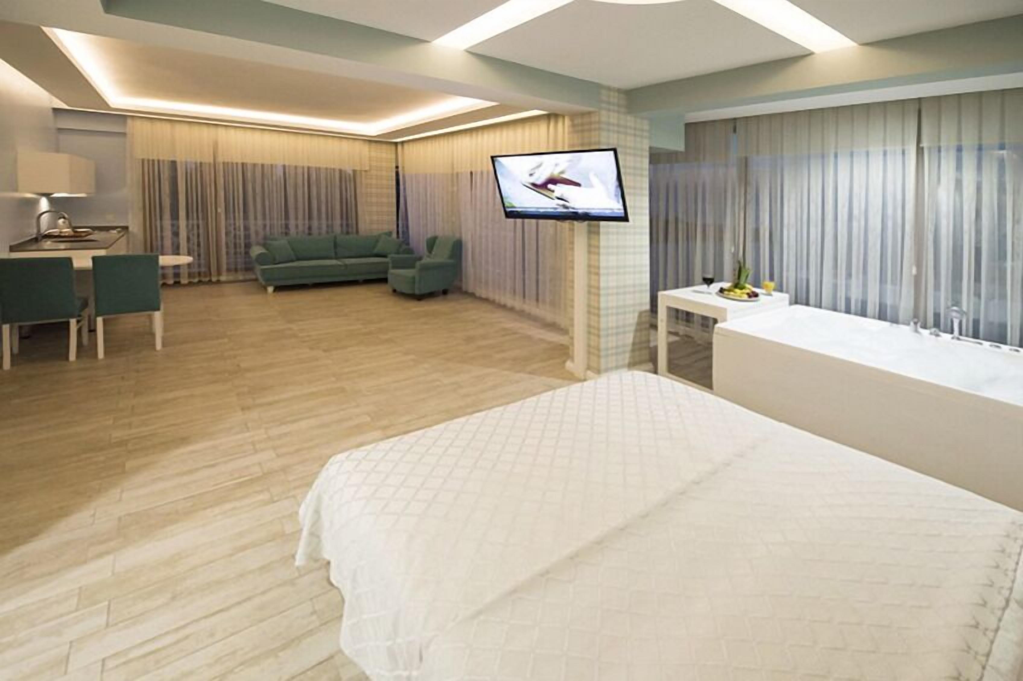 Une chambre d’hôtel moderne équipée d’un lit, d’un téléviseur fixé au mur, d’un coin salon avec un canapé et des fauteuils, et d’une petite table sur laquelle est posé un vase de fleurs.