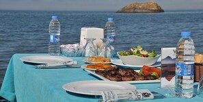 A table set with plates, cutlery, water bottles, and food items against a backdrop of the sea and a rocky island.