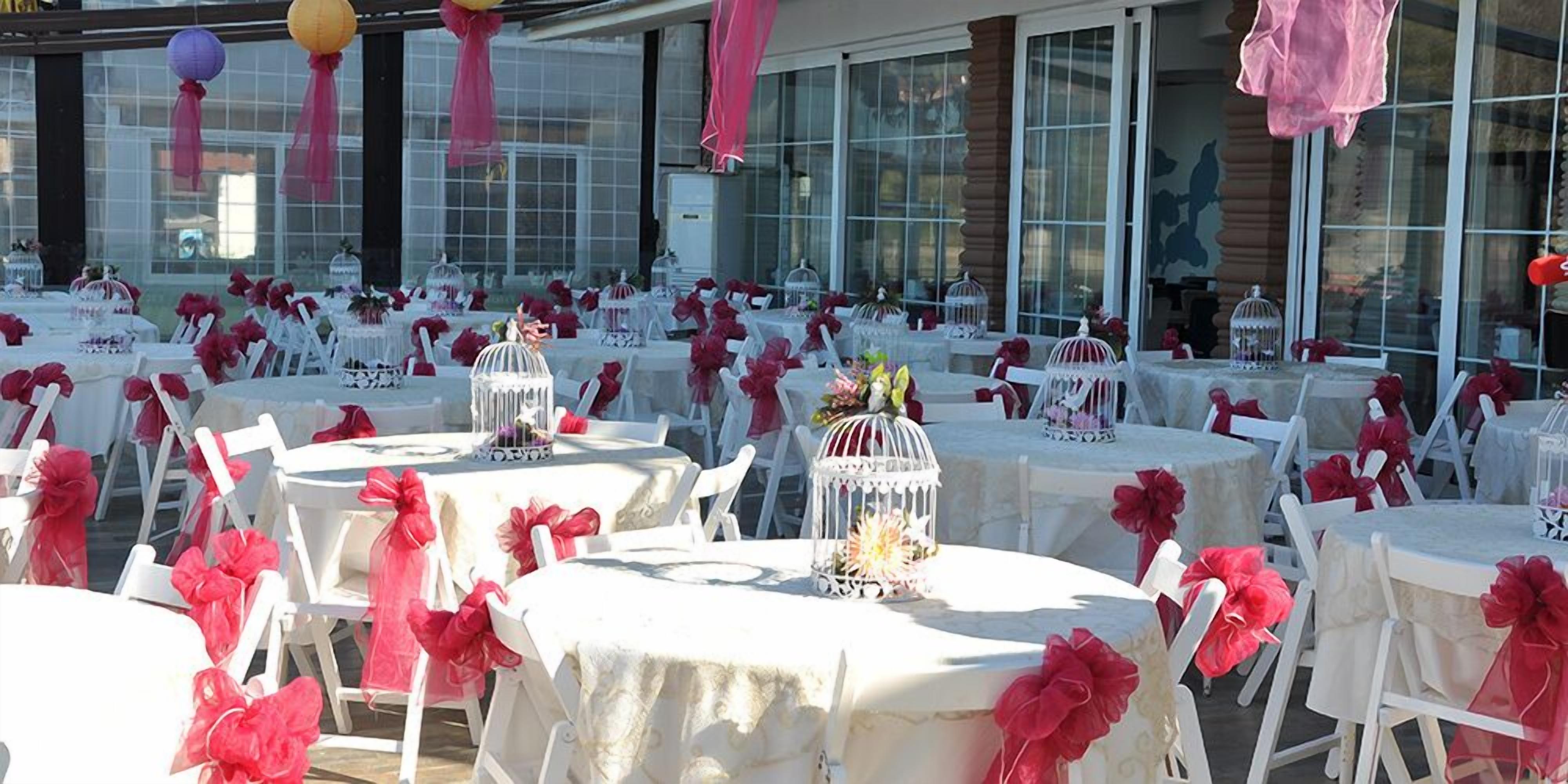 Un espacio para eventos al aire libre con mesas redondas cubiertas con manteles blancos, decorado con flores rojas y sillas blancas.