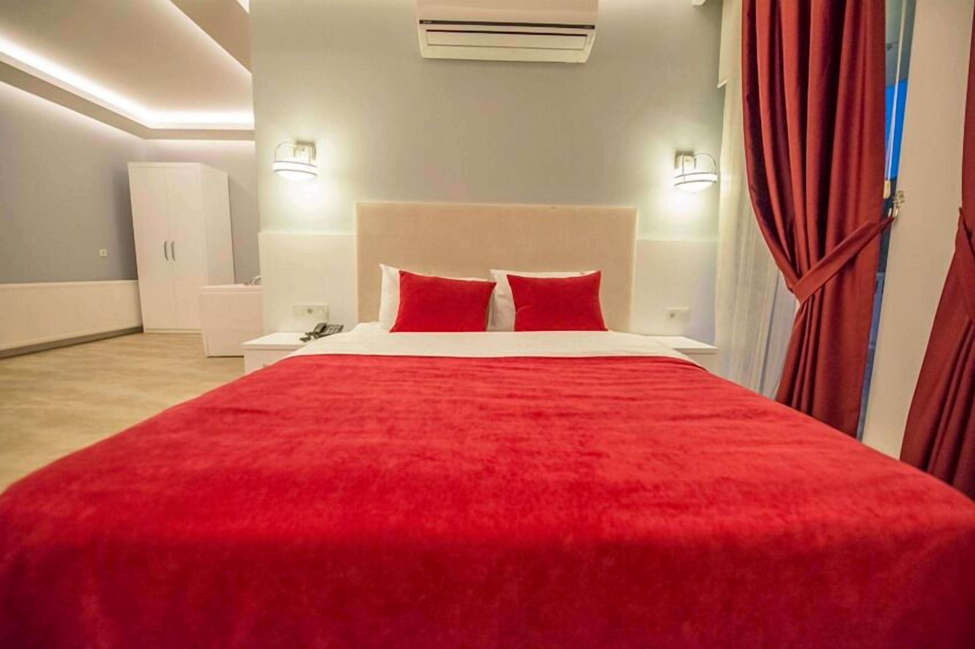 A modern hotel room with a large bed, red bedding, a white headboard, and a nightstand with a lamp.