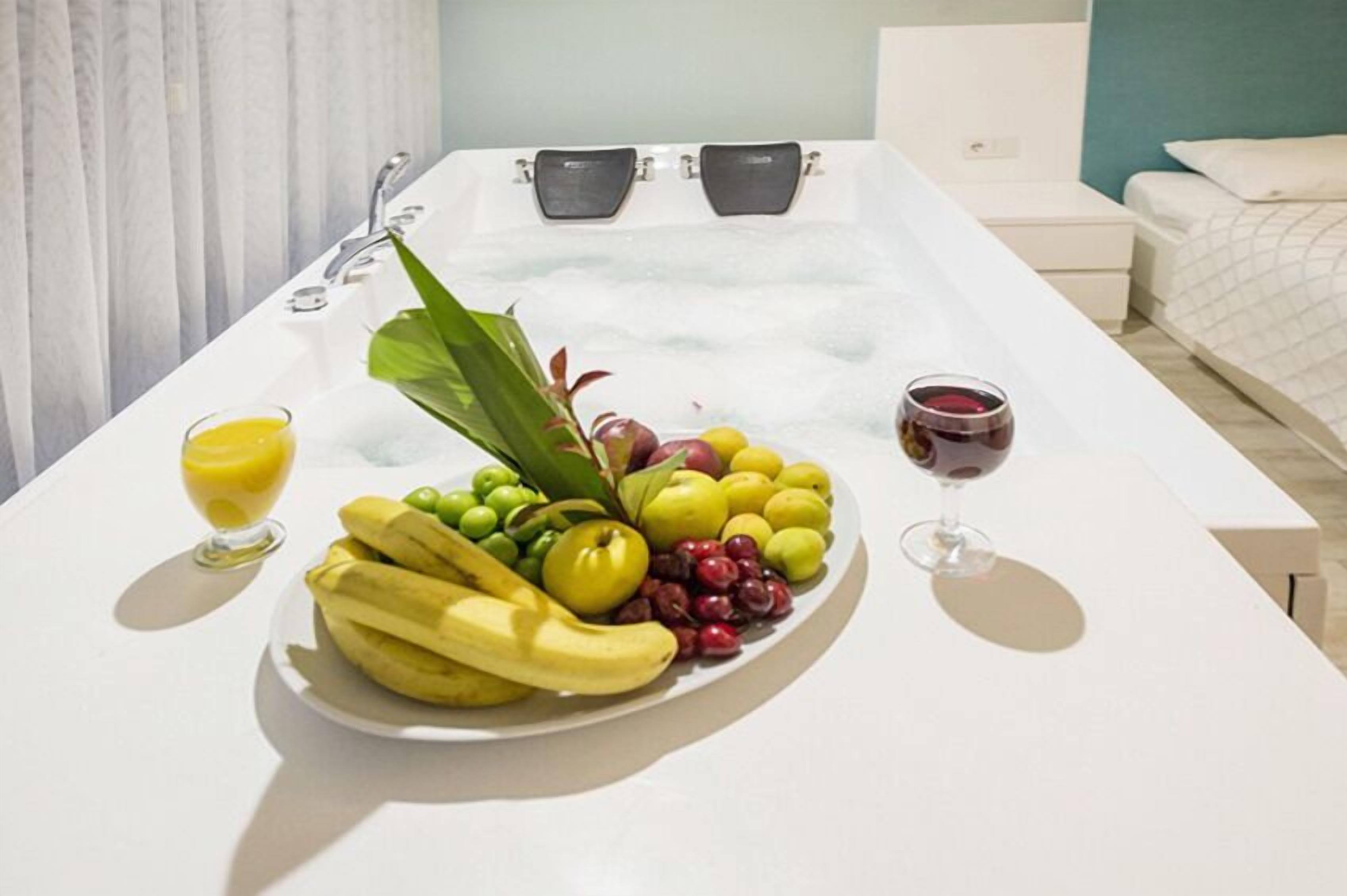 Una habitación con bañera, cama y una mesa con frutas y una copa de vino.