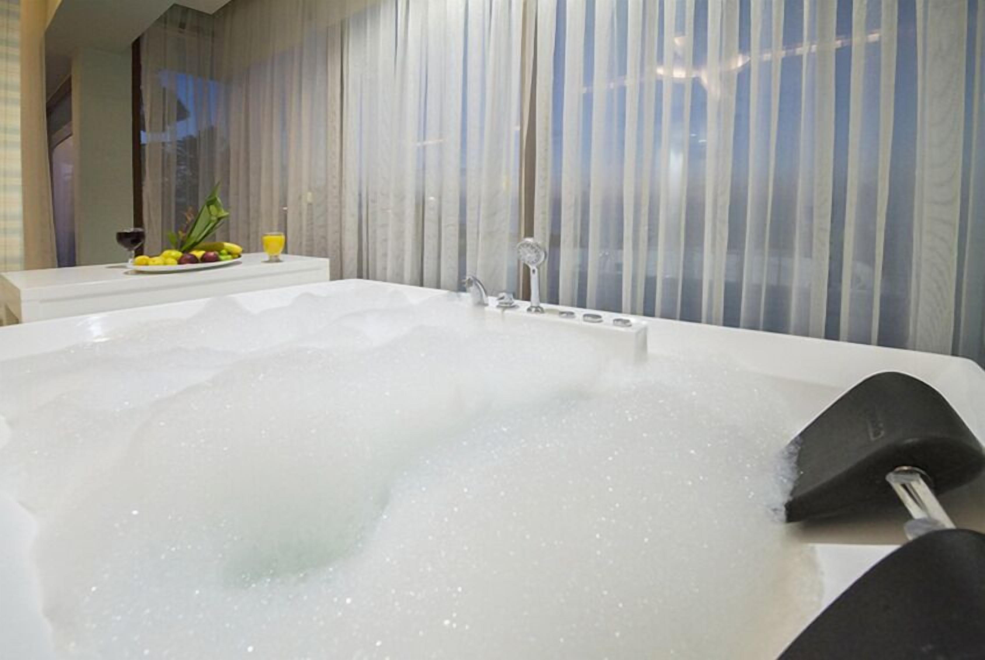 Una bañera grande llena de burbujas, una mesita con frutas y un vaso de jugo, y una ventana con cortinas.