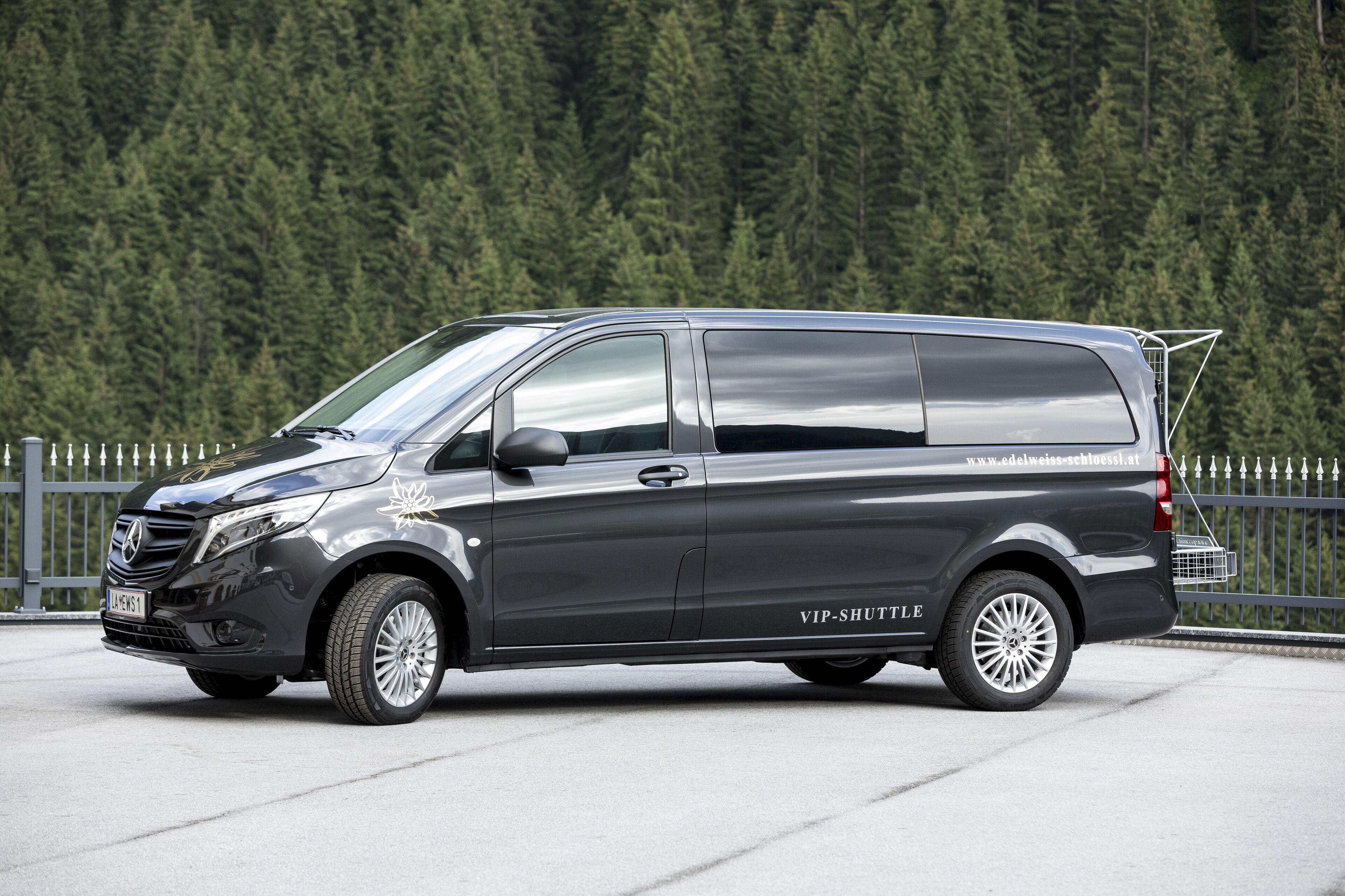 A black van with "VIP SHUTTLE" text on the side parked on a concrete surface with a metal fence and trees in the background.