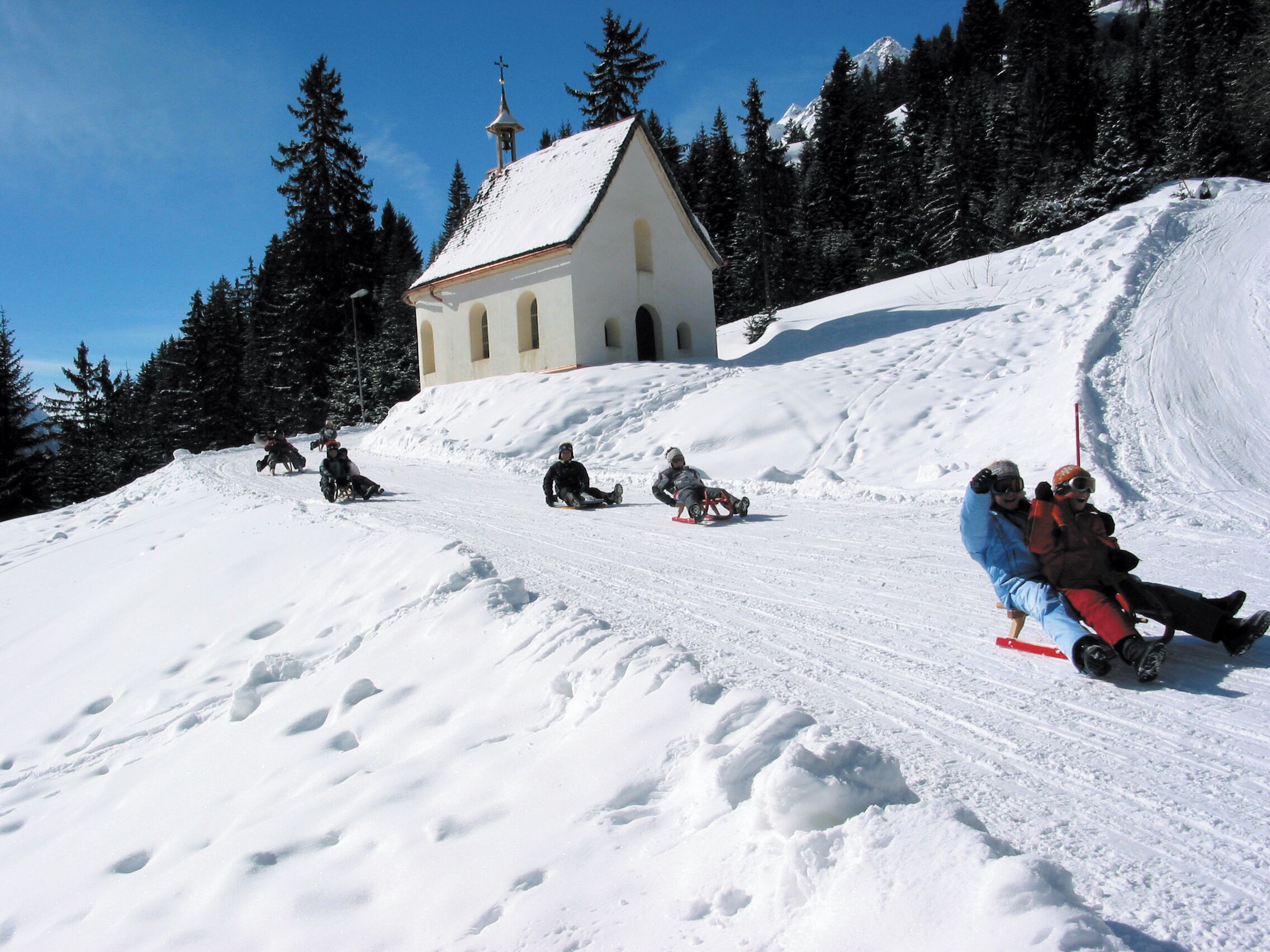 A snow-covered slope with a small white building with a bell tower, surrounded by trees.