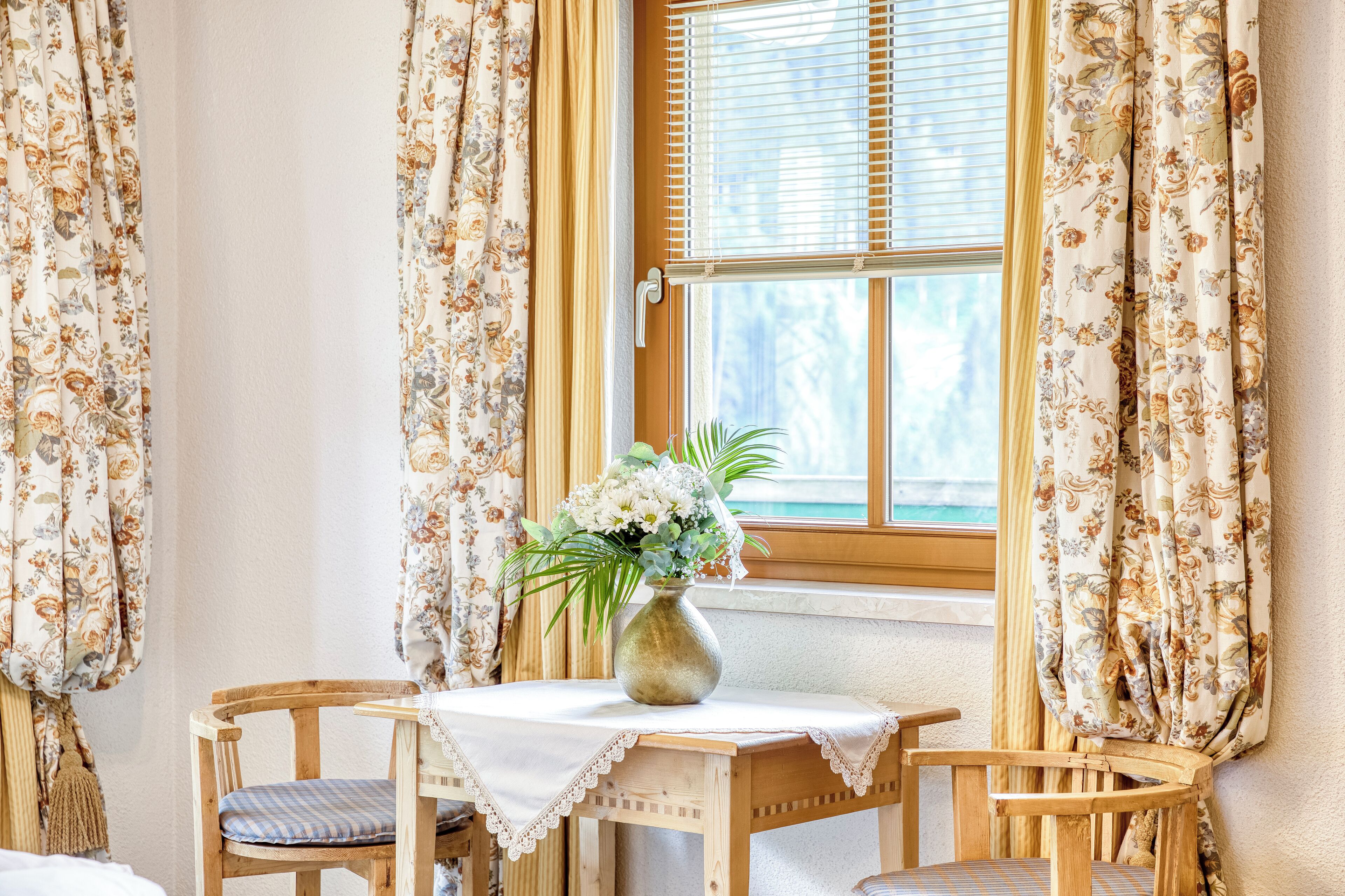 A room with a wooden table and chairs, a vase with flowers, and curtains with a floral pattern.
