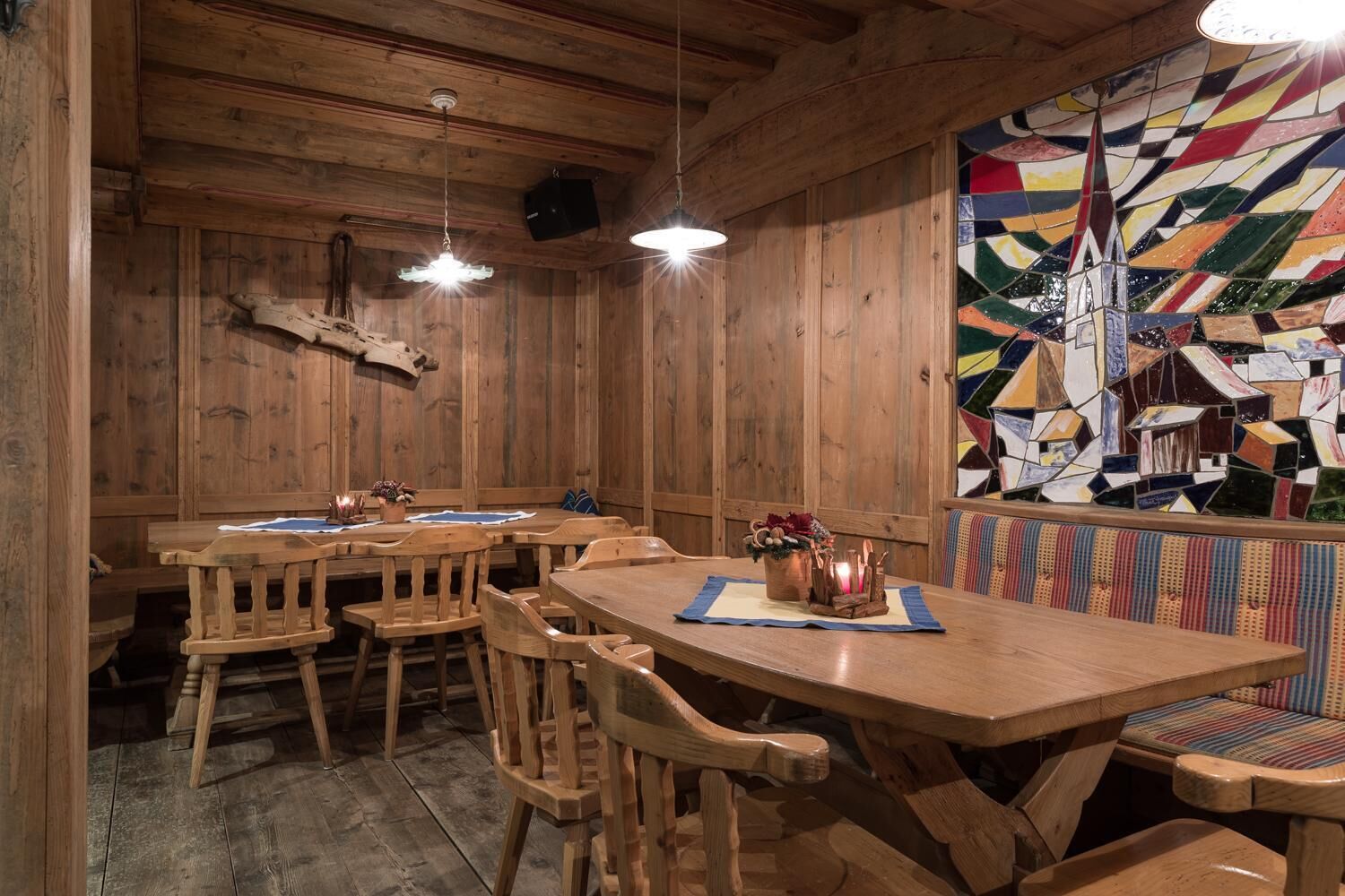 A wooden interior with a long table and benches, a mosaic wall, and wooden ceiling beams.