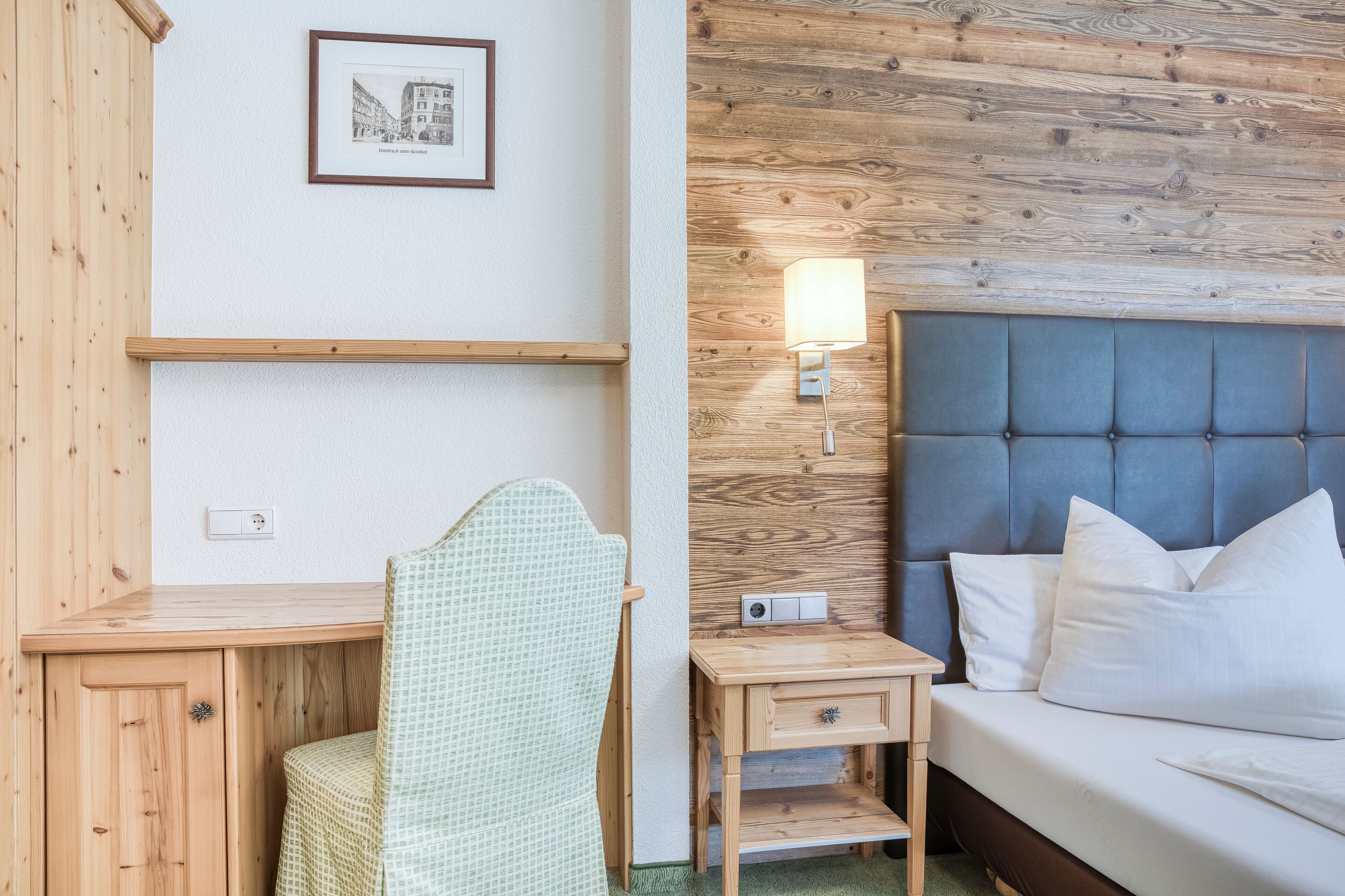 Una habitación con un escritorio de madera, una silla, una mesita de noche con lámpara, una cama con almohadas y un cuadro en la pared.