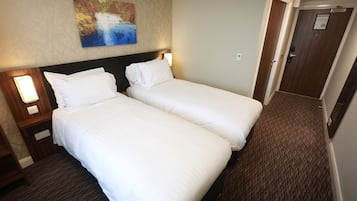 A hotel room with two single beds, a nightstand with a lamp, a painting on the wall, and a carpeted floor.