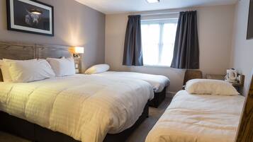A hotel room with two beds, a headboard, a window with curtains, a bedside table with a lamp, and a framed picture on the wall.
