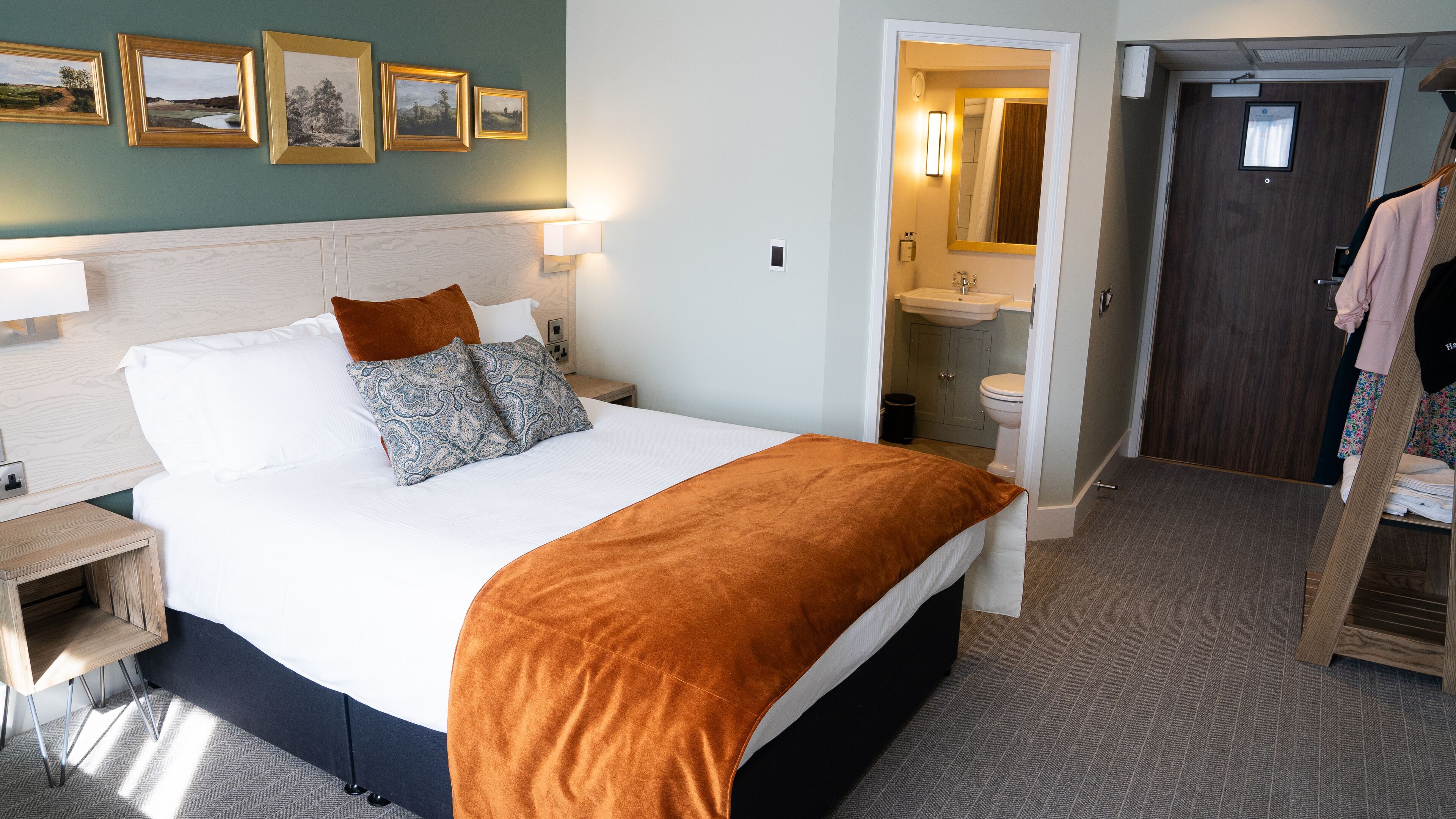 A hotel room with a large bed, a wooden nightstand, a bathroom with a sink and mirror, and a coat rack with clothes.