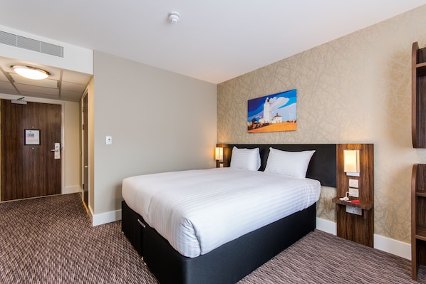 Une chambre d’hôtel avec un grand lit, une tête de lit dotée d’un luminaire, un tableau représentant un phare et une table de chevet en bois avec une lampe.