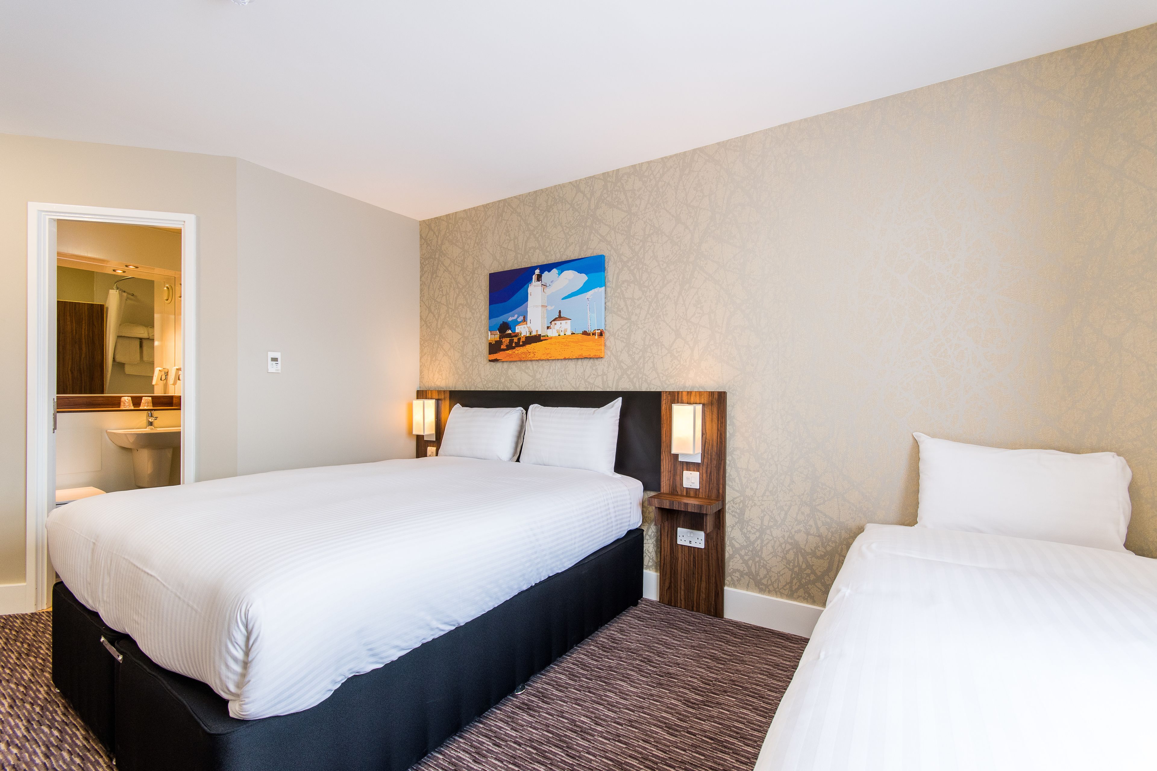 A hotel room with a large bed, a smaller bed, a painting on the wall, and a bathroom visible through an open door.