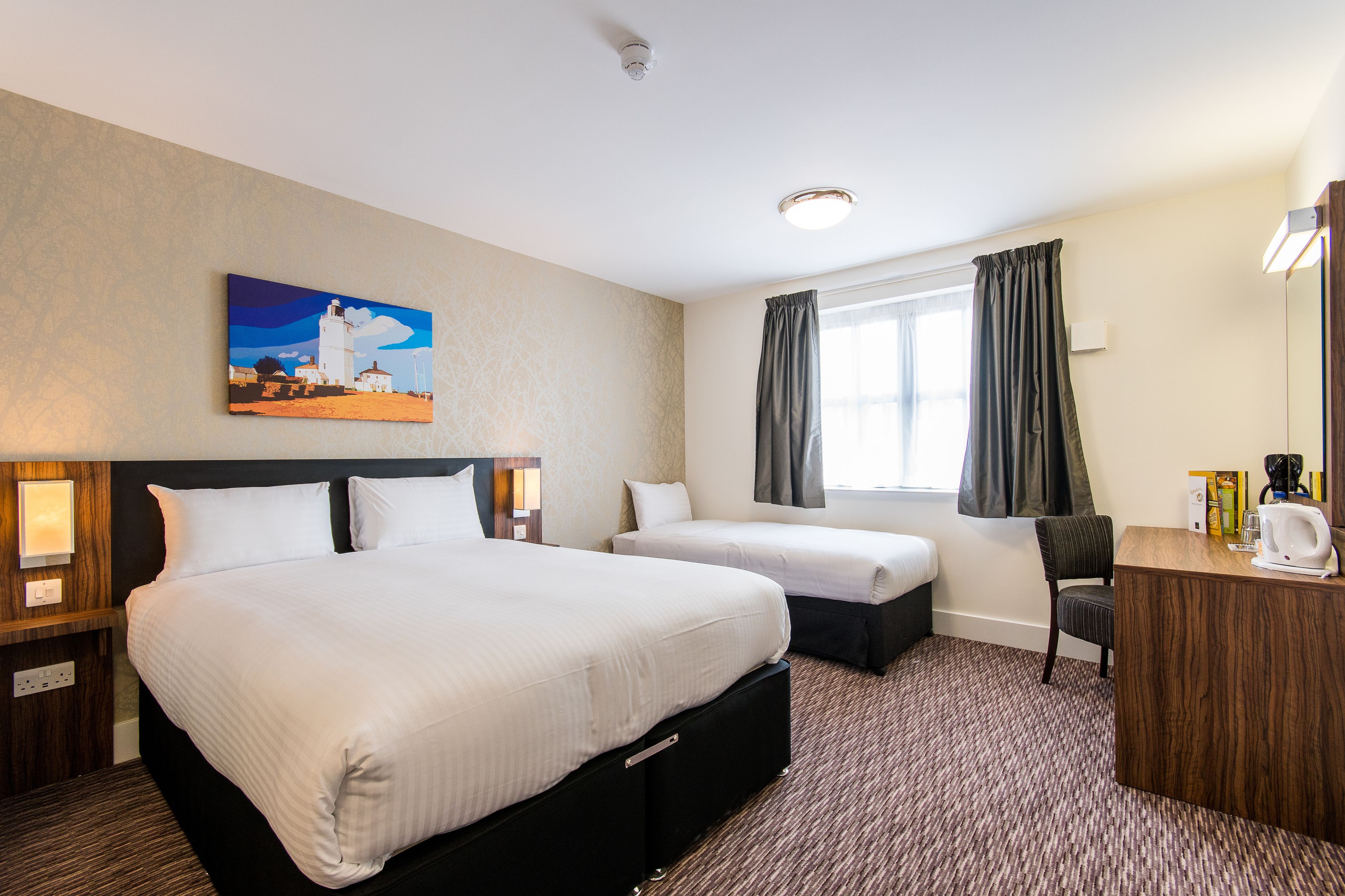 A hotel room with a large bed, a smaller bed, a desk with a chair, a painting on the wall, and a window with curtains.