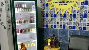 A room with a refrigerator stocked with beverages, a small table with a statue and candle, and a chair. - Pousada Axé (Salvador)