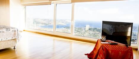 A spacious room with a large window offering a city view, a flat-screen TV, and a small table with a remote control.