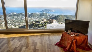A room with a large window overlooking a coastal landscape, a wooden floor, and a table with a remote control and a cup. - Viva B&B (Saronikos)